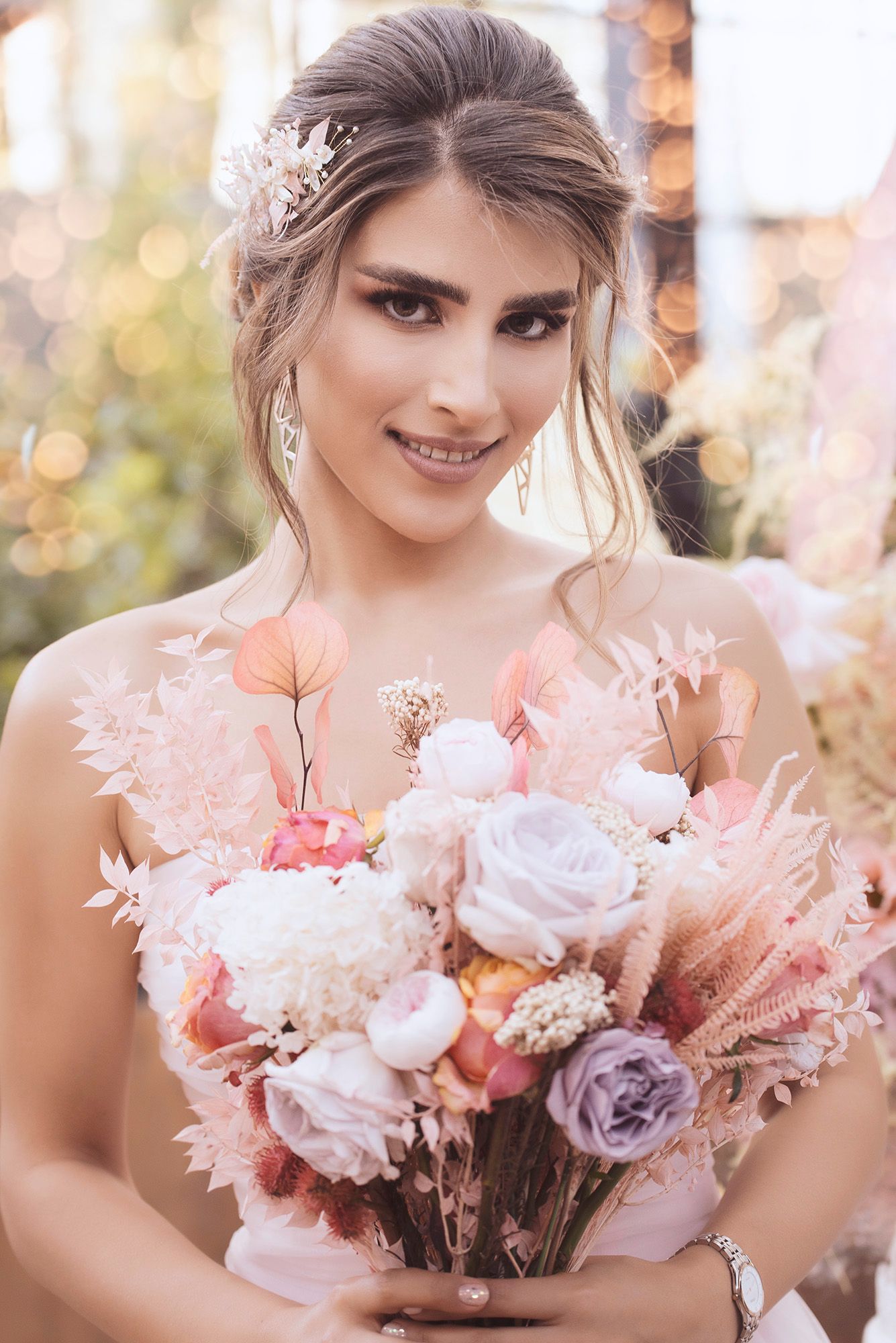Smiling bride holding a pastel bouquet of roses and dried flowers, wearing a pink dress with floral hair accessories.