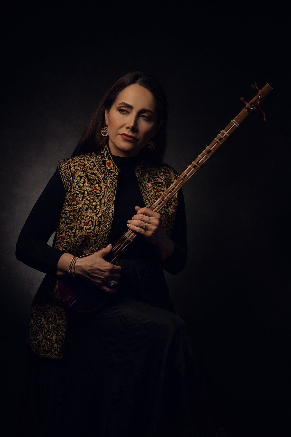 Woman in embroidered vest holding a traditional stringed instrument in dark studio portrait.