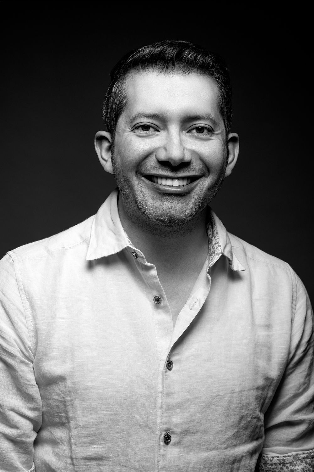 Black and white portrait of a smiling man in a white linen shirt against a dark background.