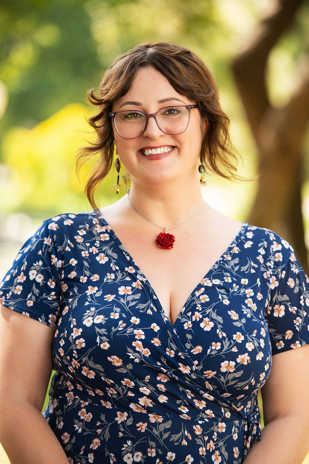 Smiling woman in blue floral dress with rose necklace, outdoor professional portrait.