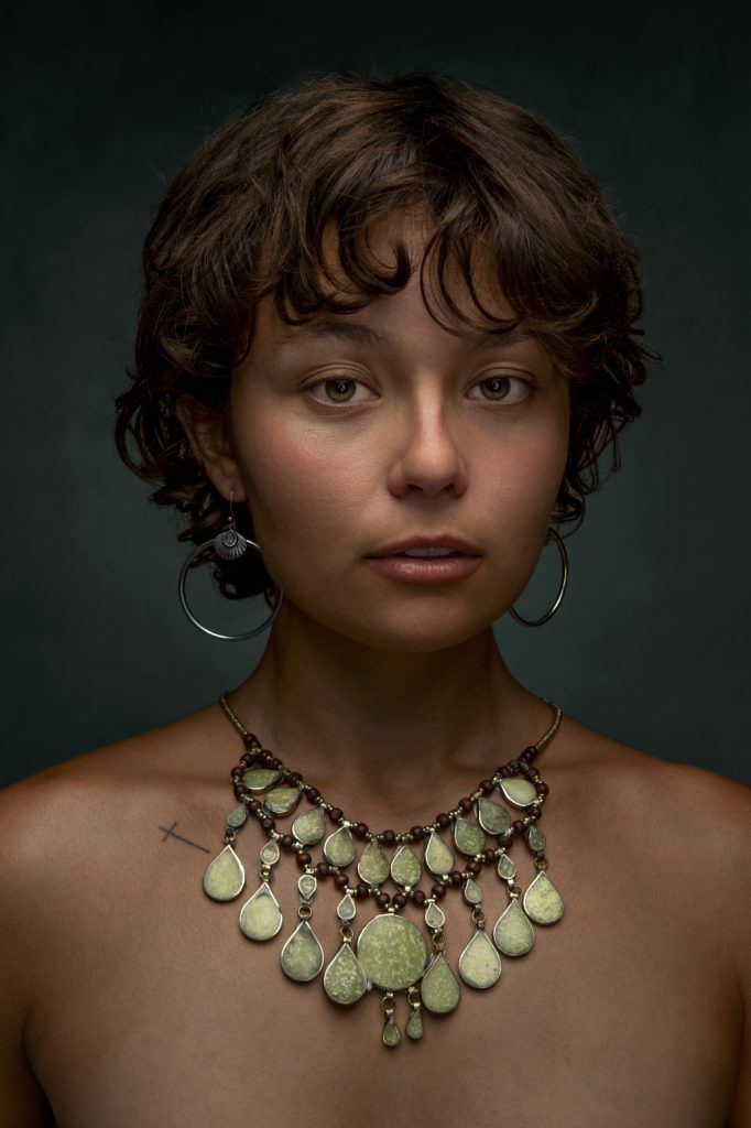 Studio portrait of a young woman with short curly hair wearing a green stone necklace and hoop earrings.