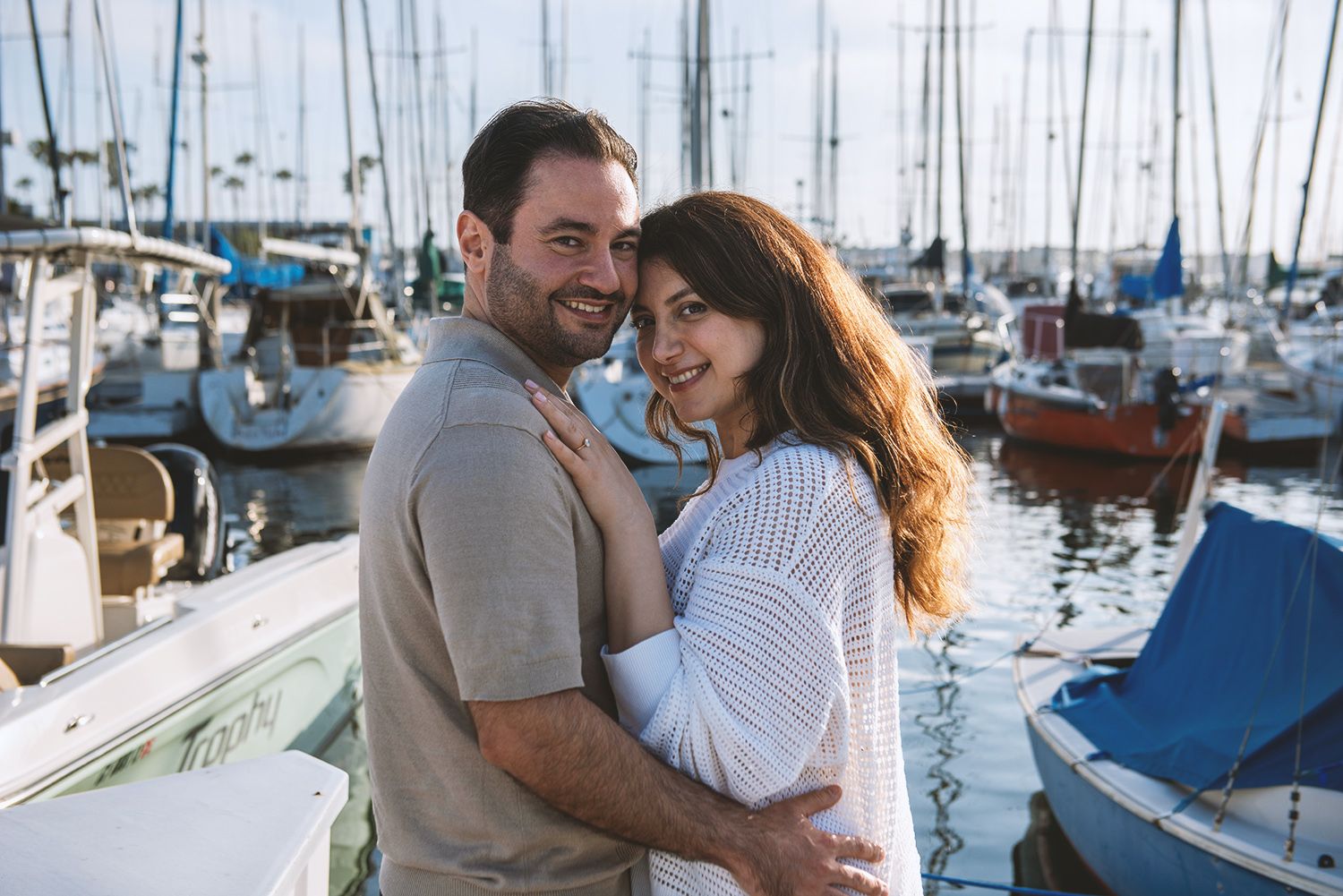 Happy couple embracing and smiling at a sunny marina with sailboats in the background.