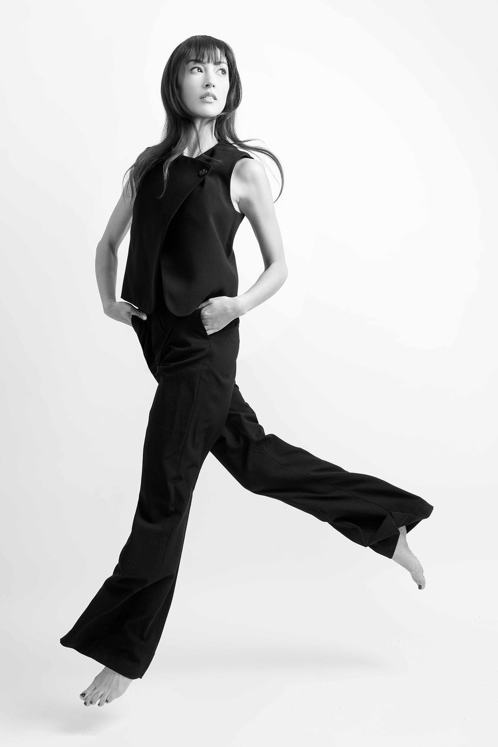 Black and white photo of model in sleeveless black pantsuit jumping gracefully barefoot.