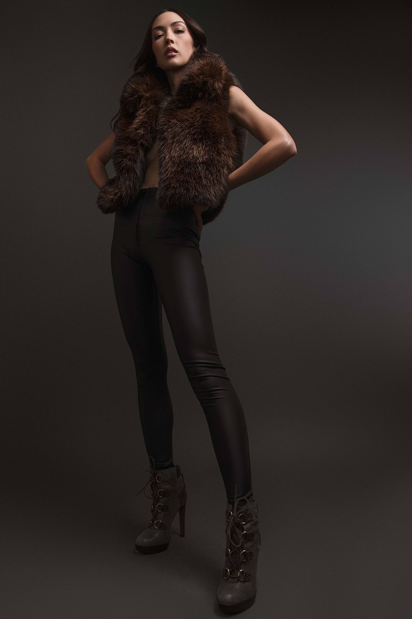 A fashion model in a brown fur vest and black leather leggings poses in high-heeled ankle boots.