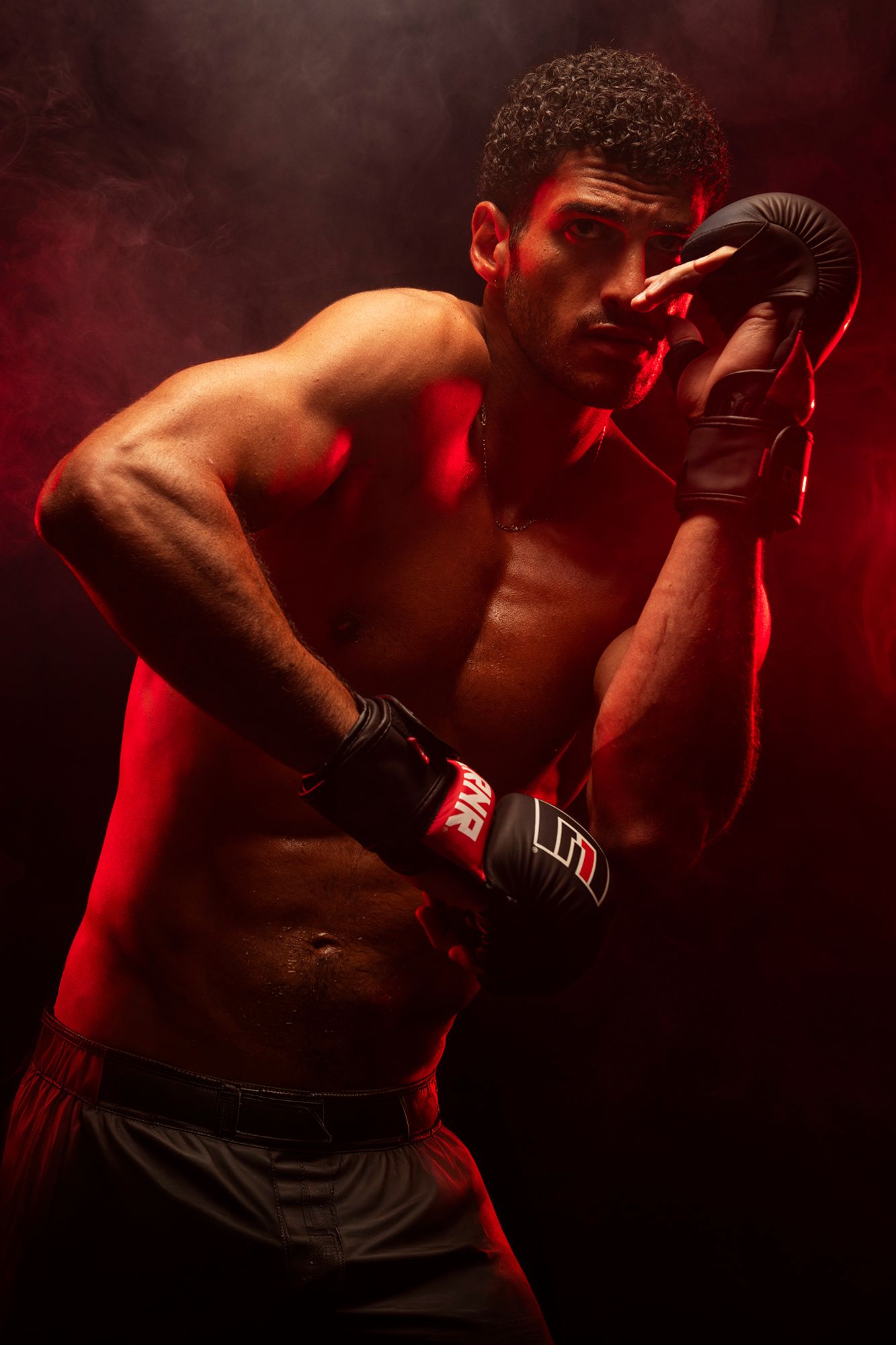 Muscular shirtless male boxer in fighting stance wearing black gloves, dramatic red smoke background.