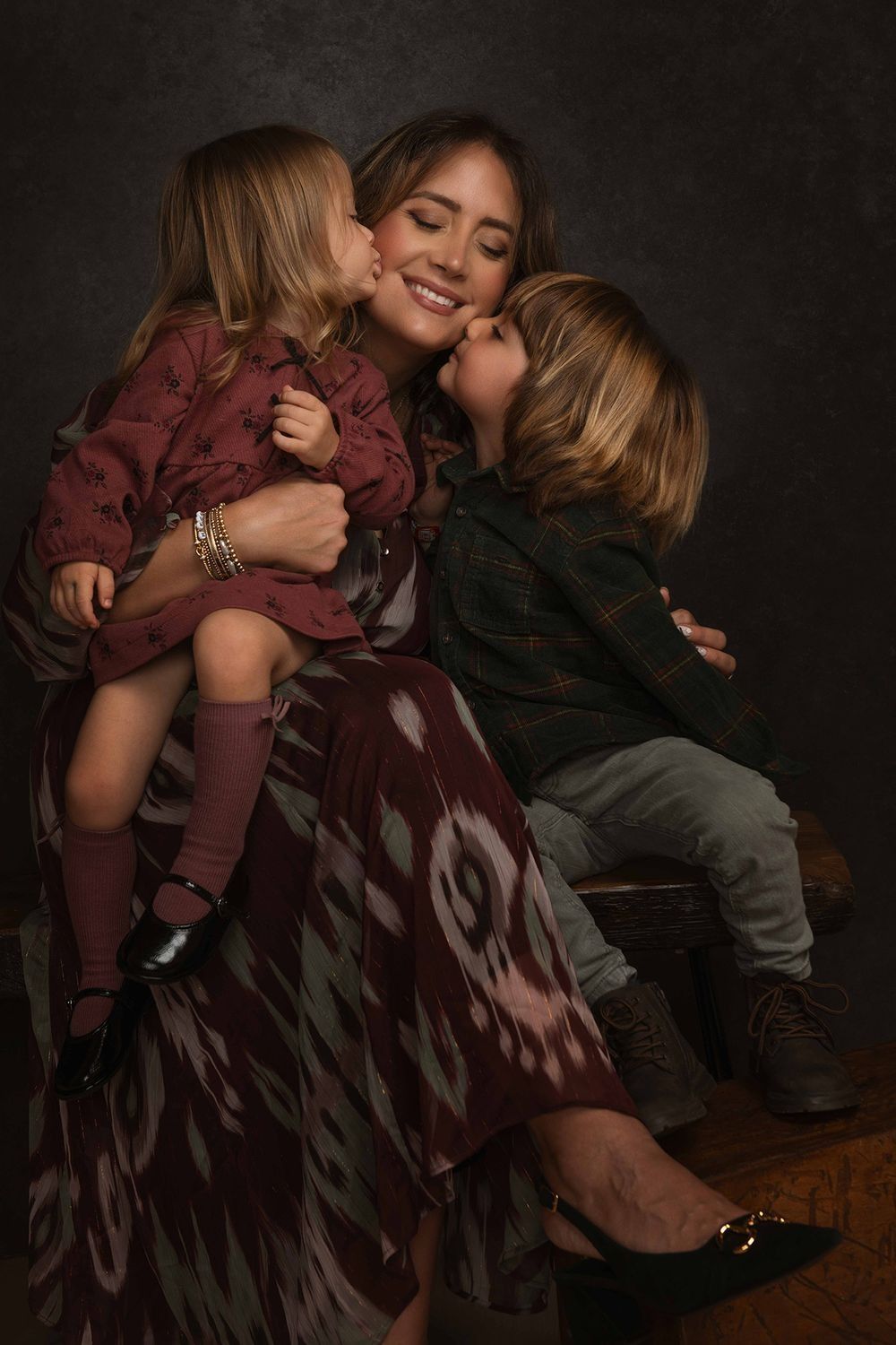 A smiling woman in a patterned maroon skirt sits holding two young children who kiss her cheeks in a warm studio family portrait.