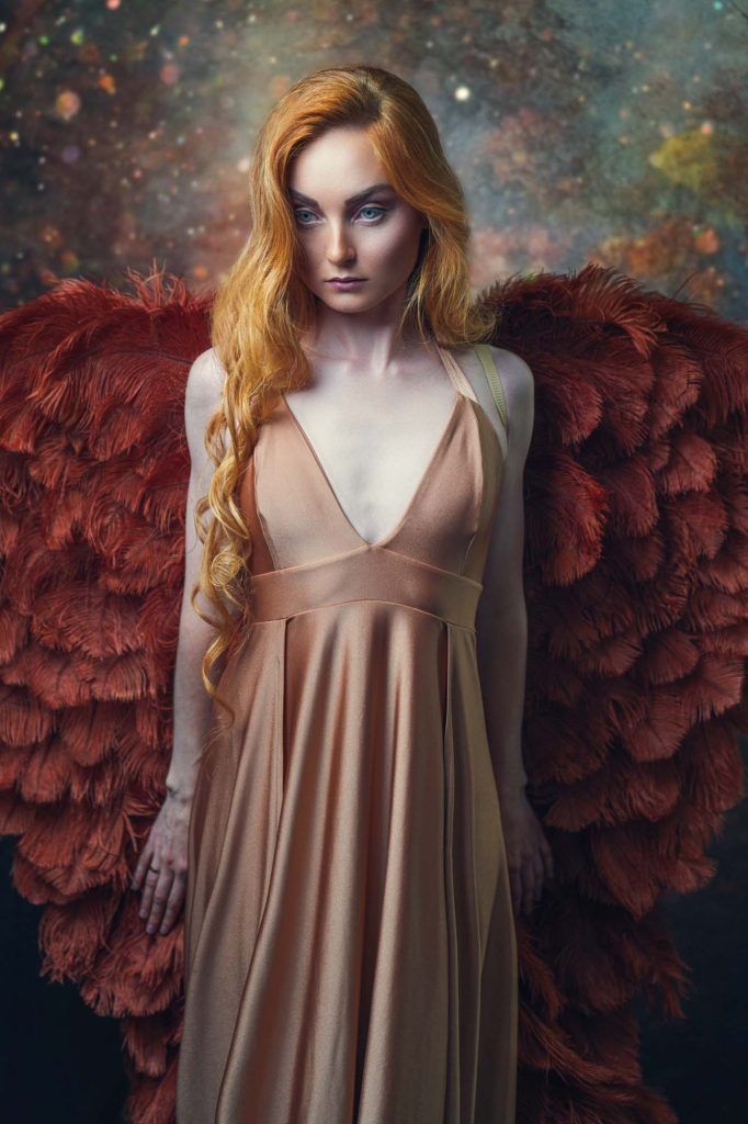 Fantasy portrait of a red-haired woman in a beige gown with large dark red feathered angel wings.