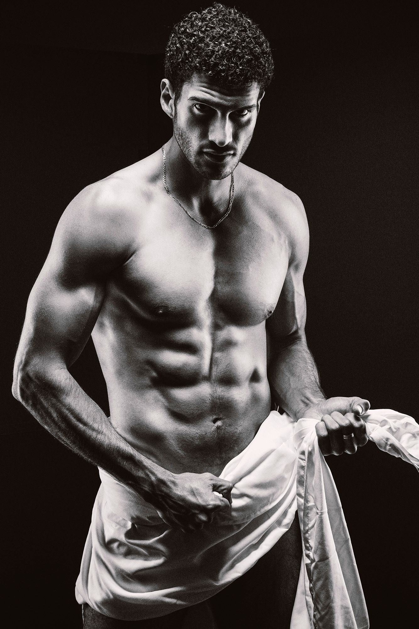 Fit shirtless man with curly hair gripping white cloth, intense gaze, black and white studio photo.