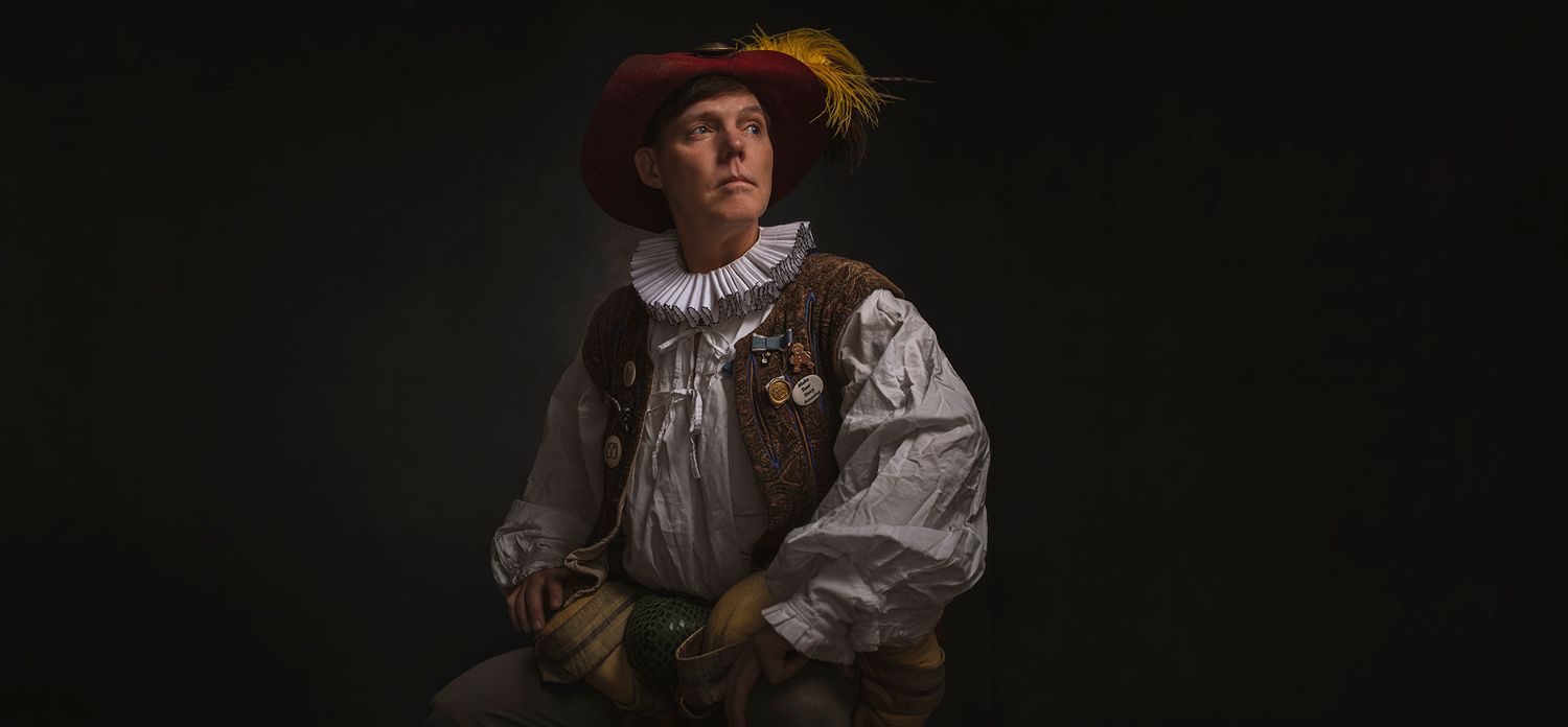 Person in historical Renaissance outfit with red feathered hat posing on dark background.