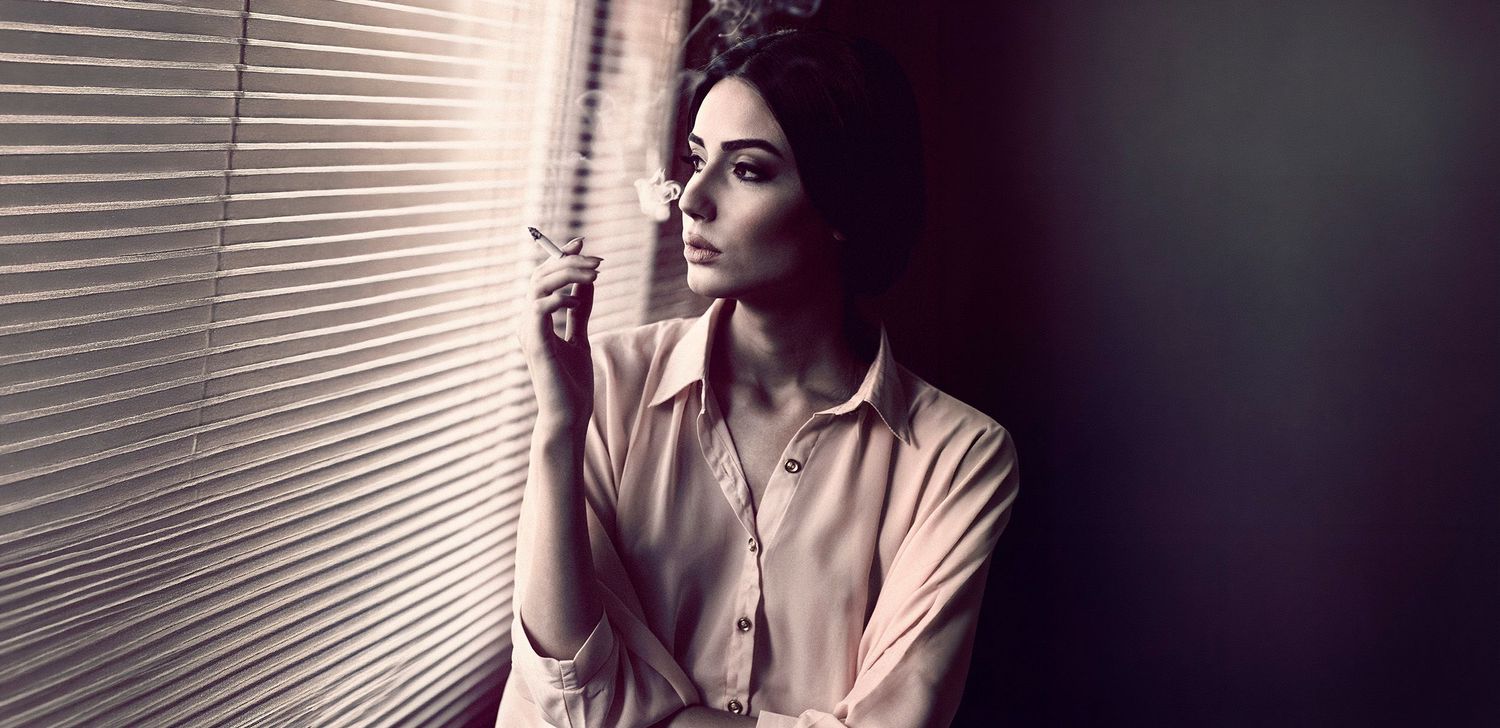 Woman in beige shirt looking through window blinds in dramatic black and white cinematic portrait.