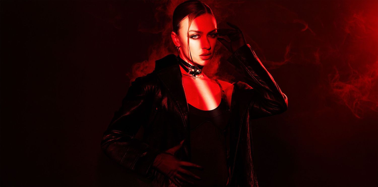 Woman in black leather jacket posed dramatically under red lighting with smoky background.