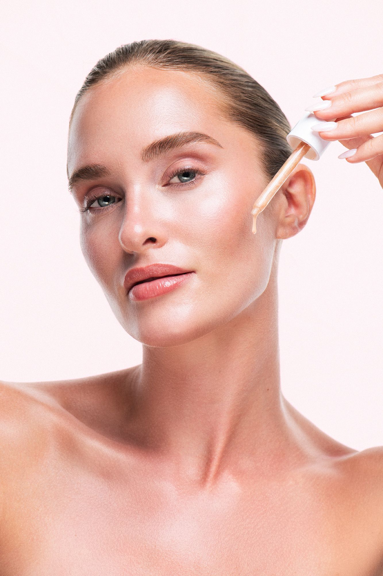 Woman applying liquid foundation serum with dropper applicator on glowing, flawless skin against light pink background.