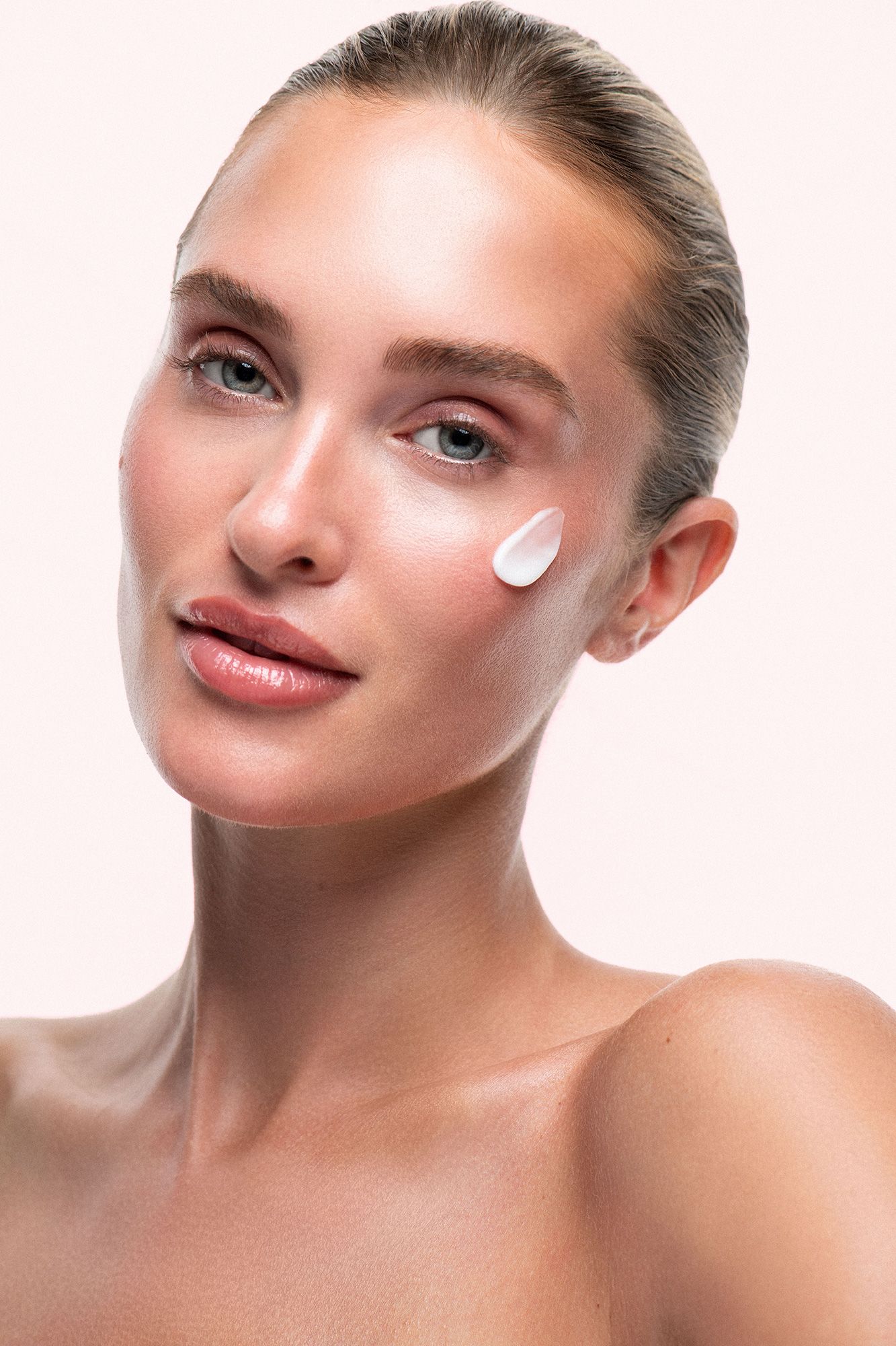 Young woman with glowing skin applying white face cream on cheek, beauty skincare concept on light background.