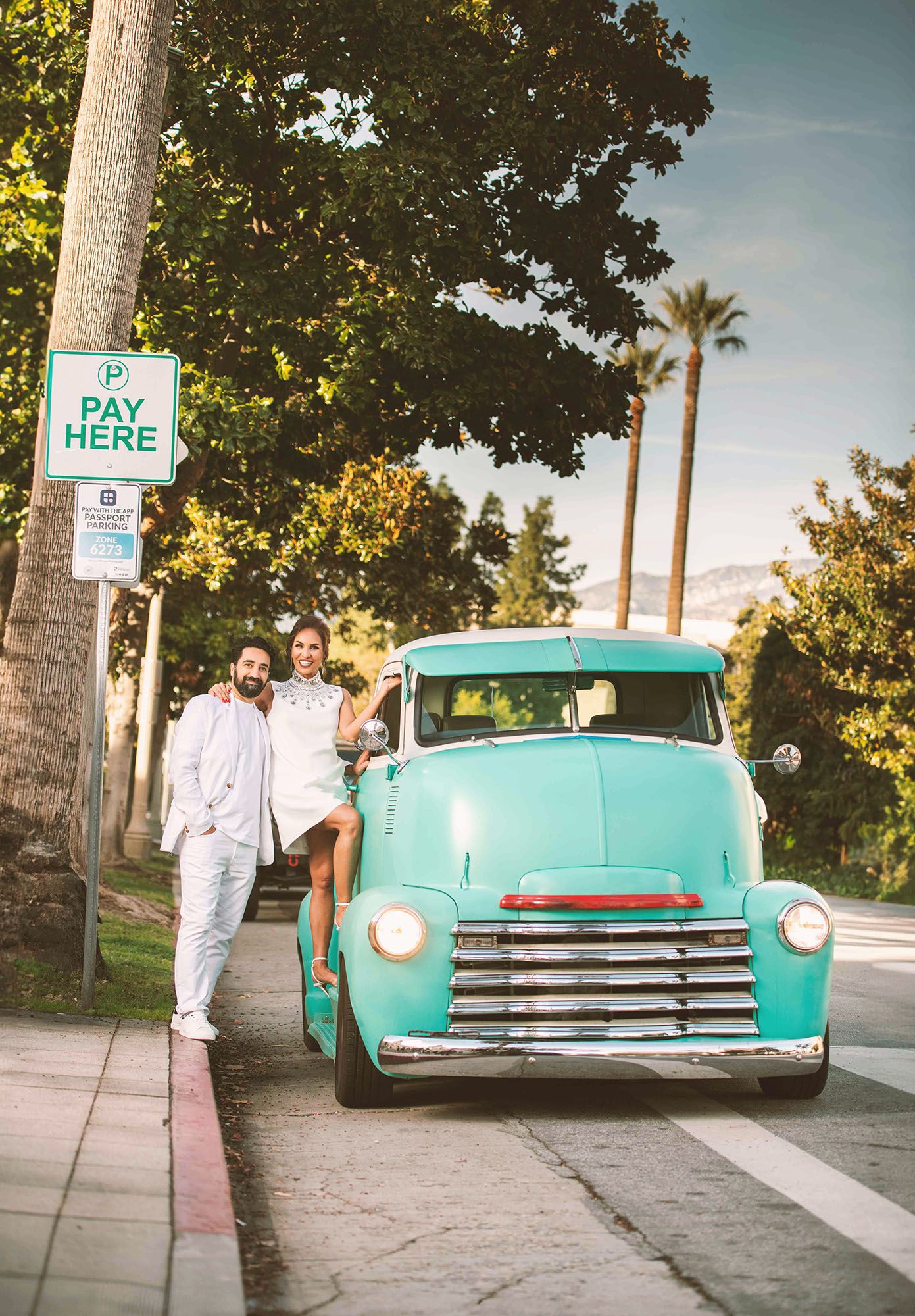 Couple posing with vintage turquoise Chevrolet truck on palm tree-lined street on sunny day.
