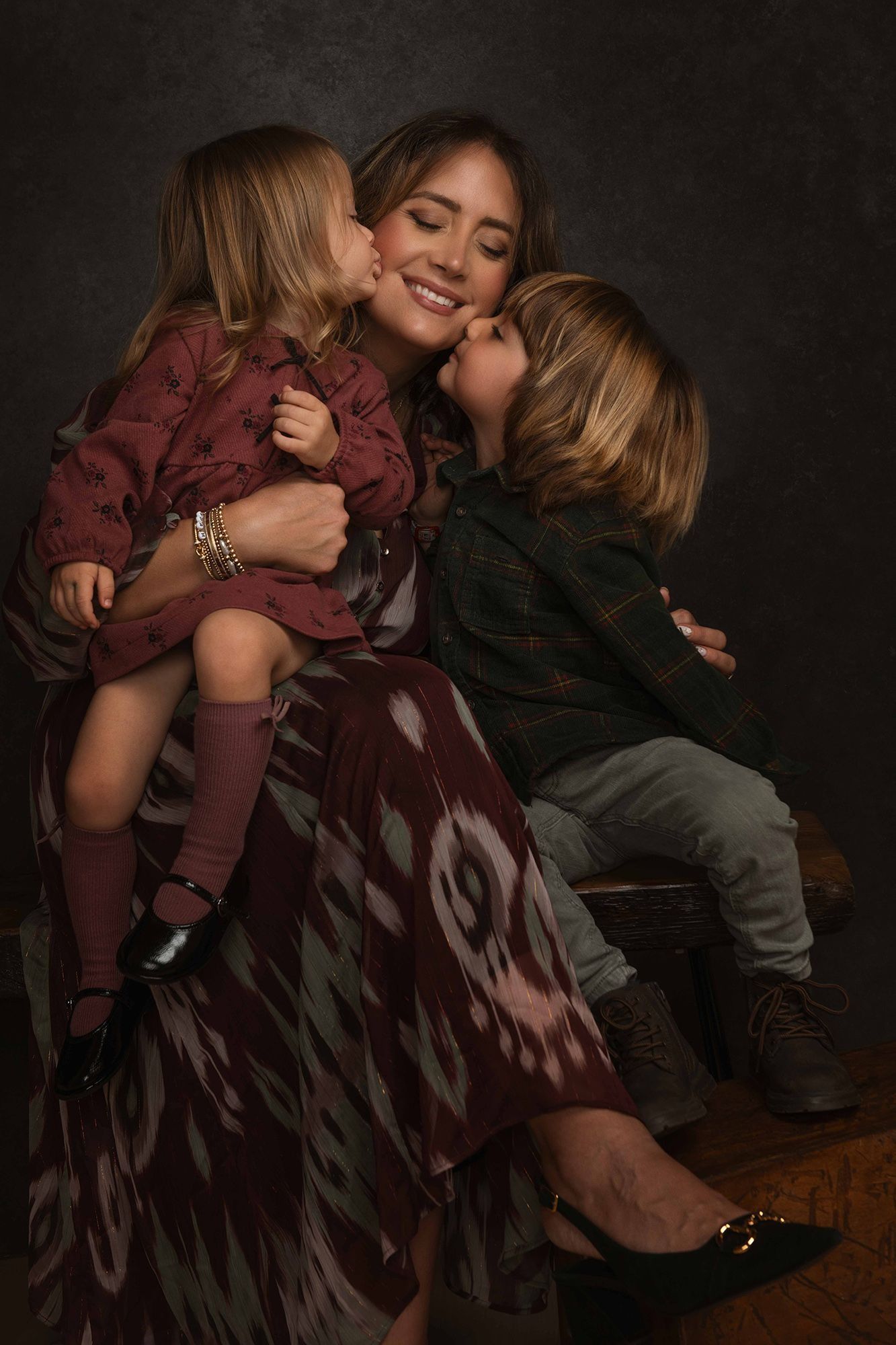 Mother smiling as two young children kiss her cheeks in a studio portrait.