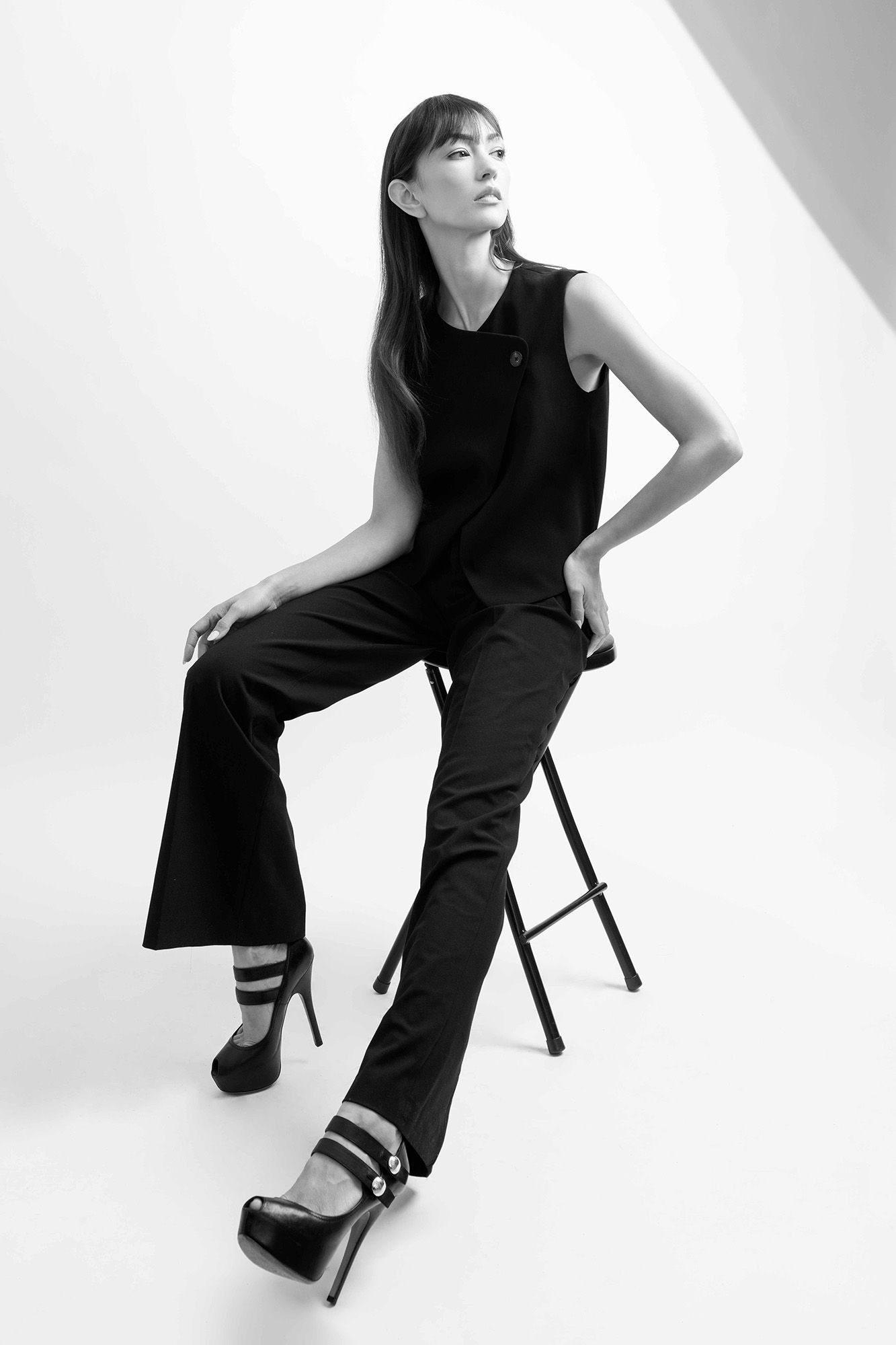 Black and white photo of a female model in black wide-leg pants and heels seated on a stool.