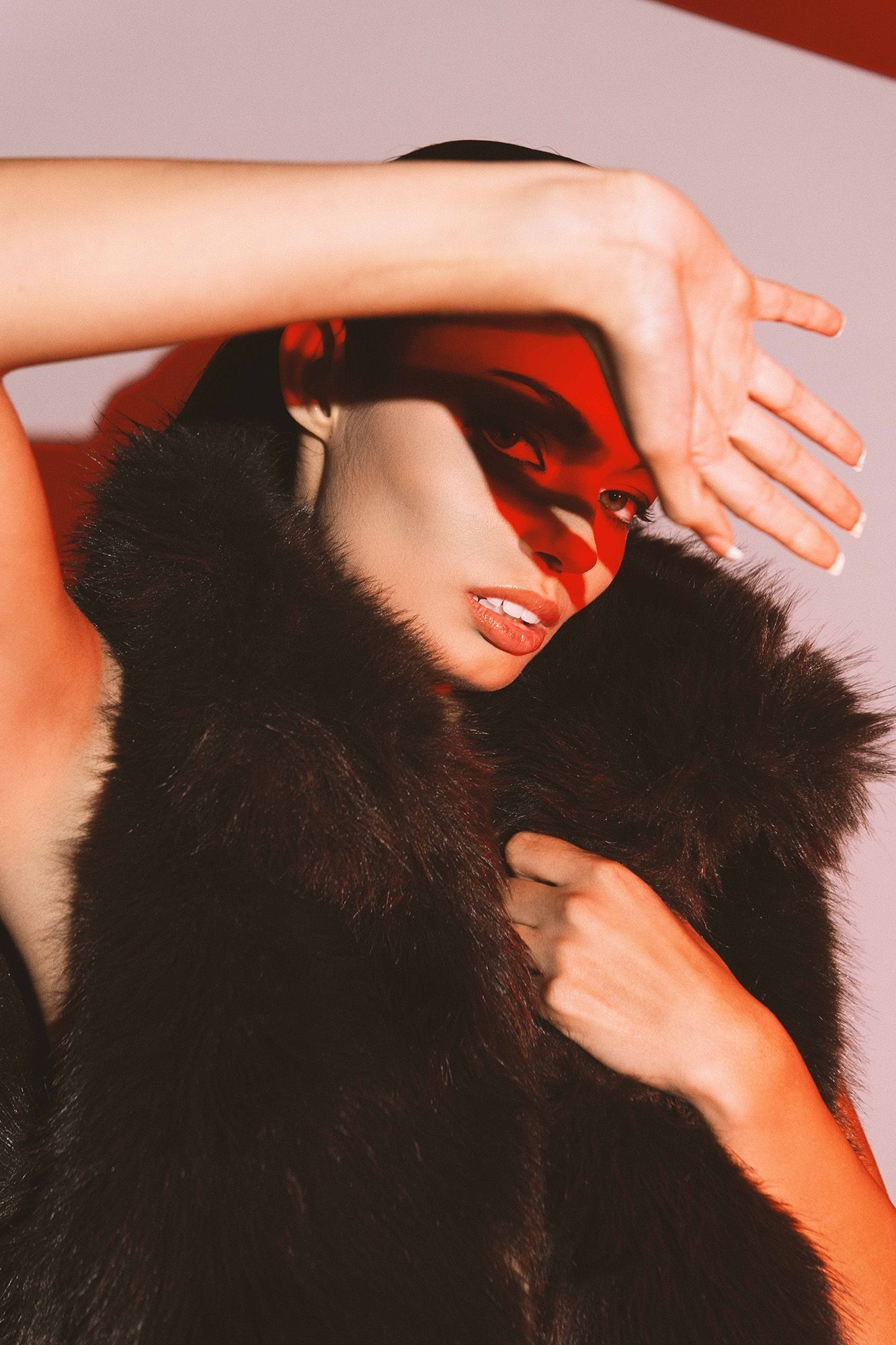 Fashion model in dark fur vest with arm raised under red dramatic studio lighting.
