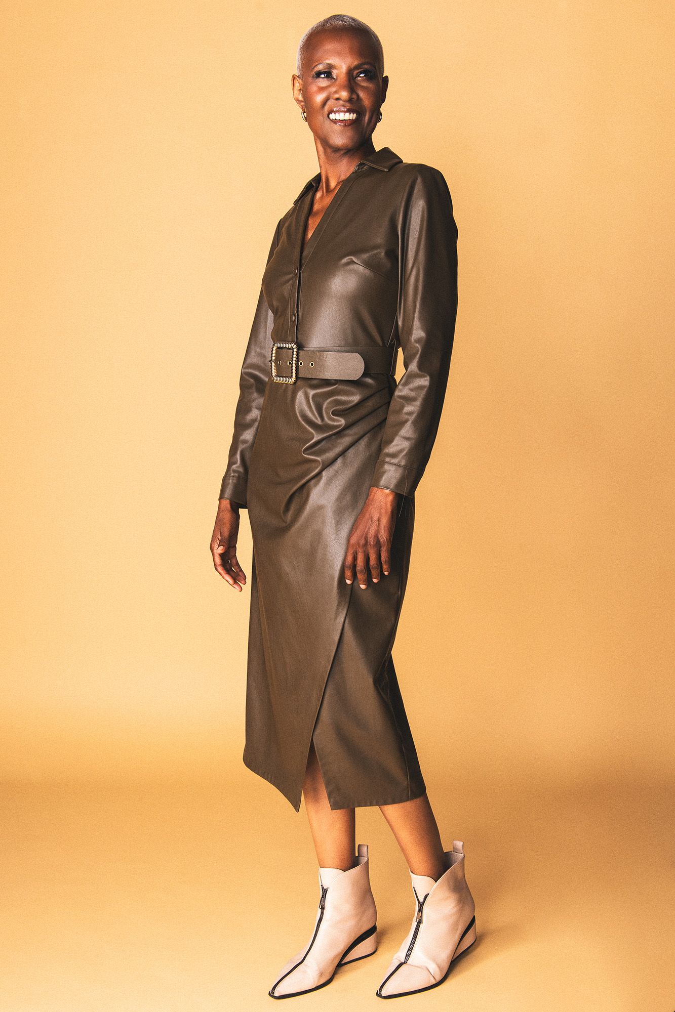 Woman in olive faux leather belted midi dress with white ankle boots, smiling against a warm beige background.
