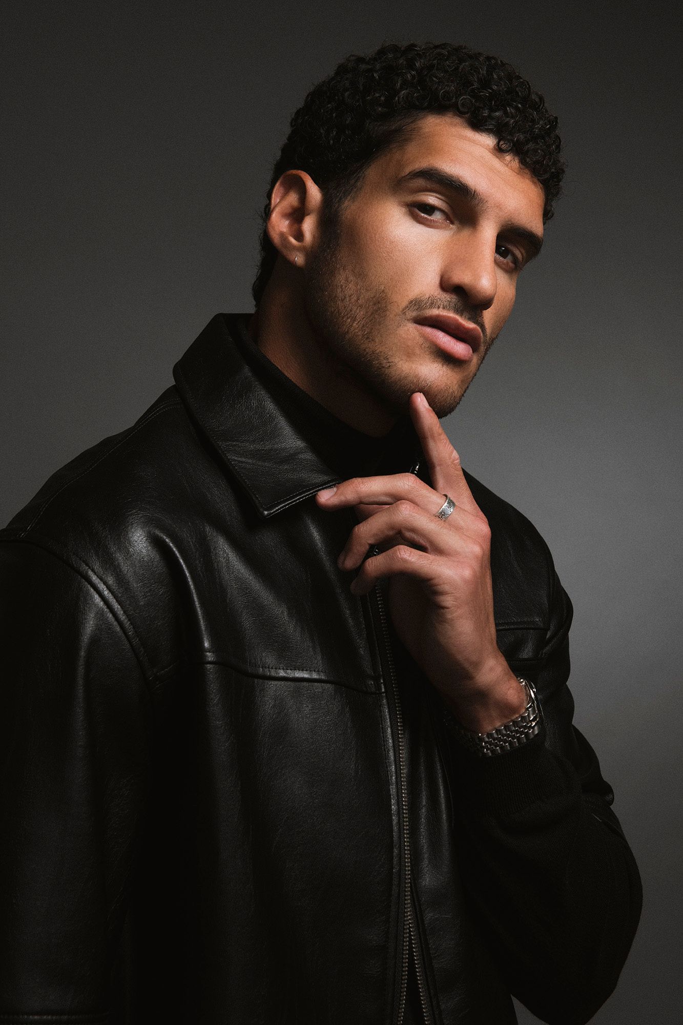 Young man in black leather jacket touches chin thoughtfully while posing against gray studio background.
