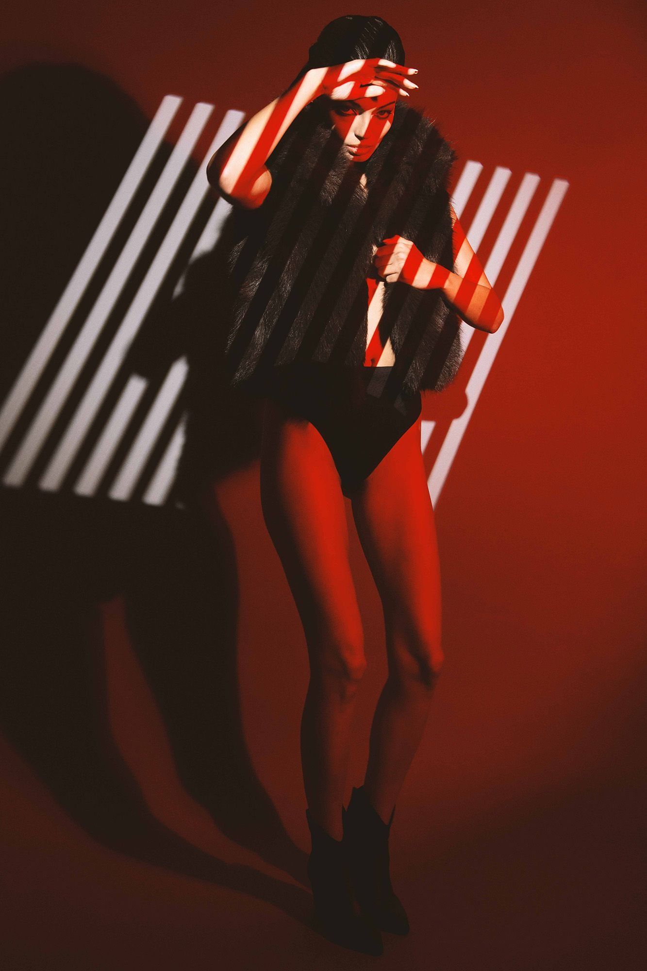 Woman in fur vest and red lighting with striped shadow pattern posing against red background.
