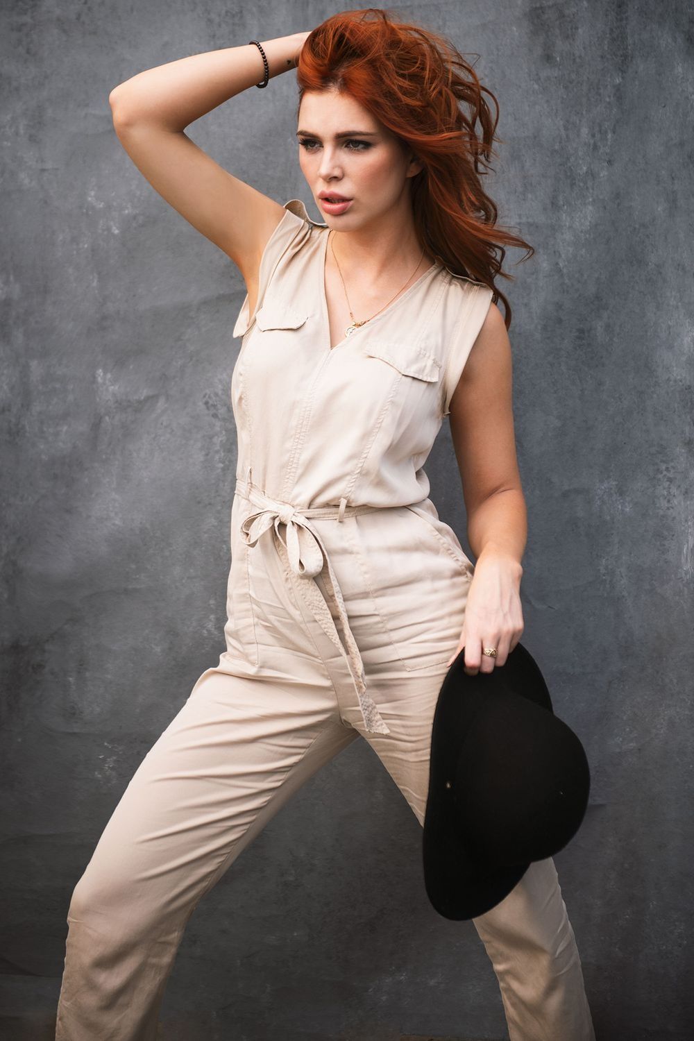 Stylish redhead woman in beige sleeveless jumpsuit holding black cap against gray textured wall.