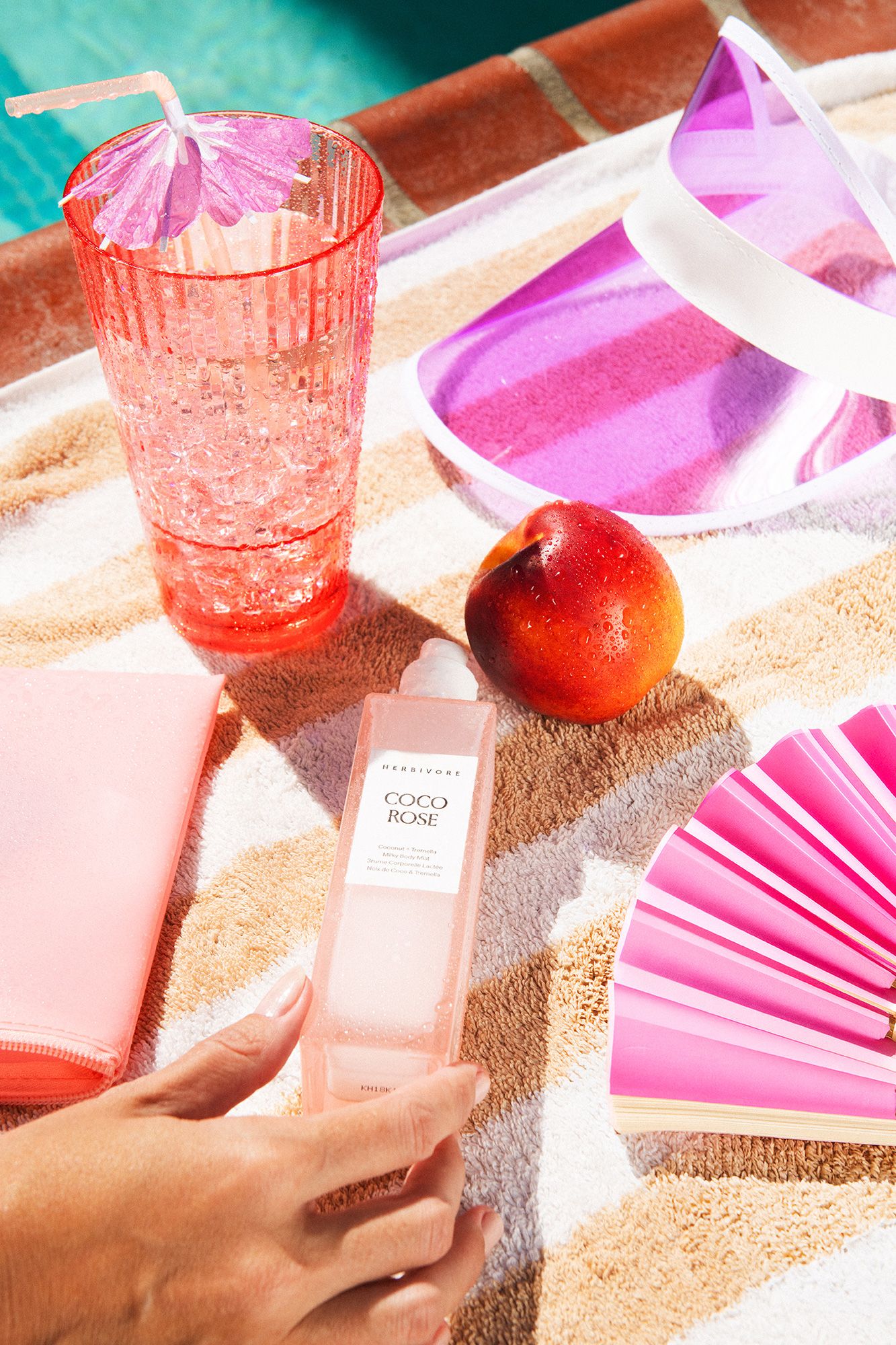 Poolside flat lay with Coco Rose body mist, pink drink, nectarine, visor, and fan on striped towel.