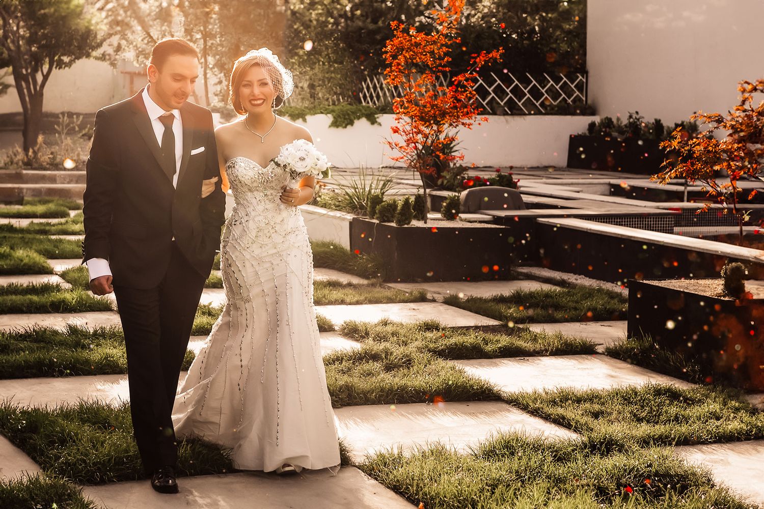 Elegant bride in beaded gown and groom in black tuxedo walking together in a sunlit garden on their wedding day.
