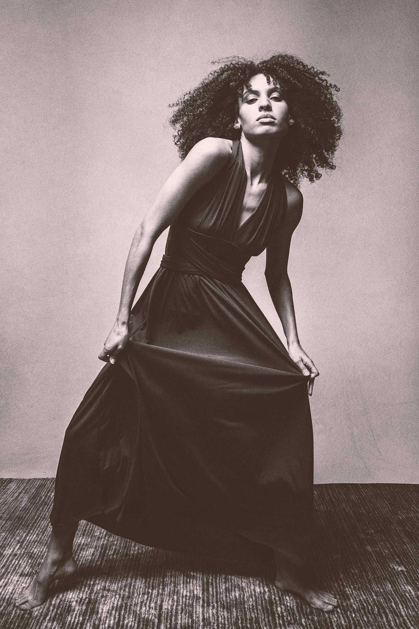 Black and white photo of a woman with curly hair in a flowing dark dress, posing dynamically in a studio.