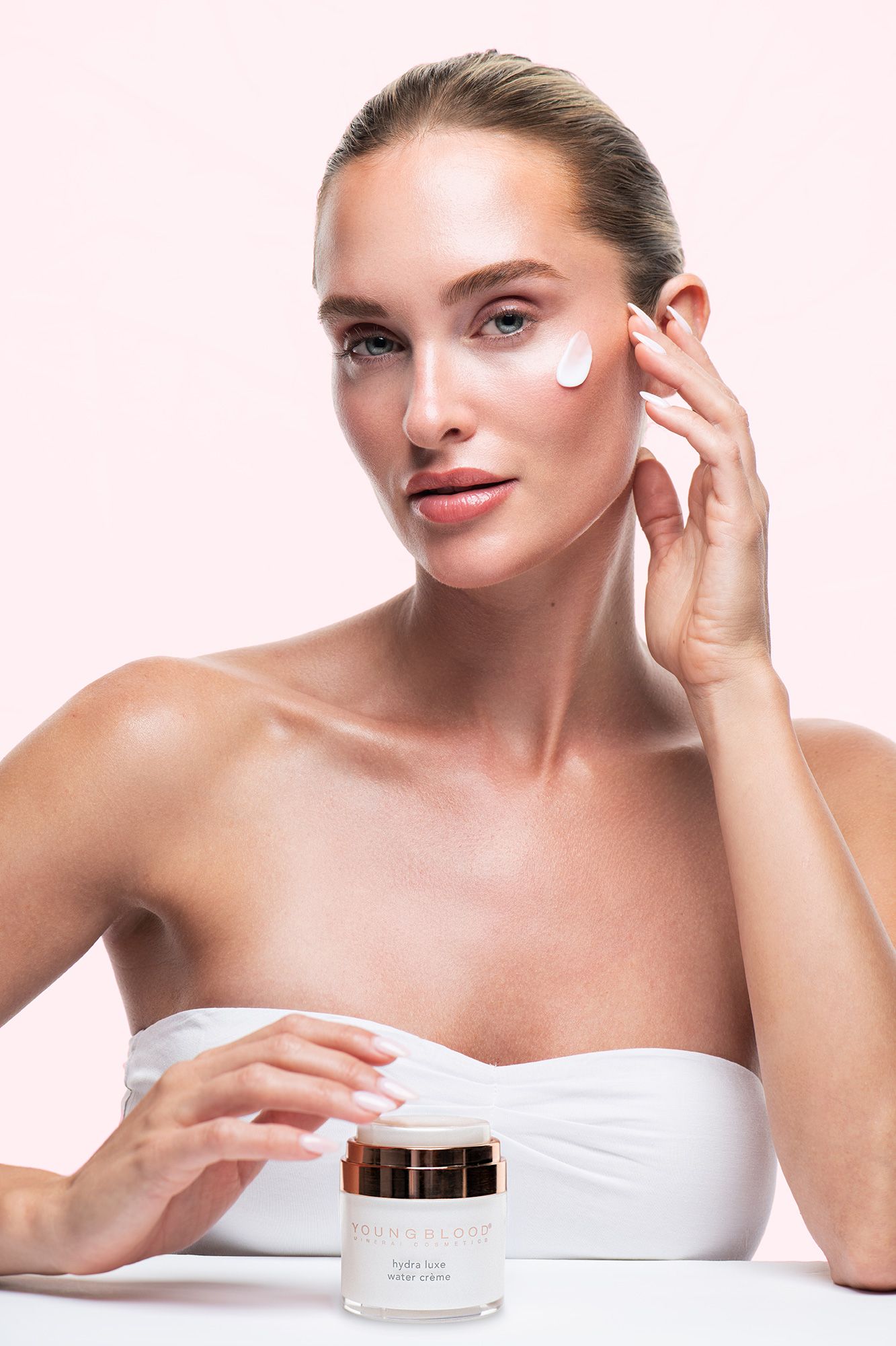 Woman applying white face cream to cheek, holding skincare moisturizer jar, beauty product advertisement on pink background.