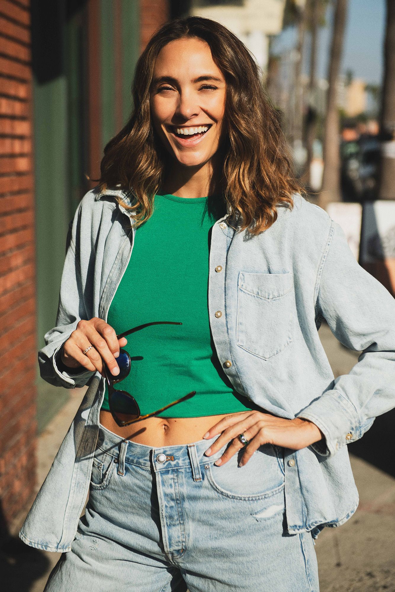 Smiling woman in green crop top, denim shirt, and light wash jeans holding sunglasses outdoors.