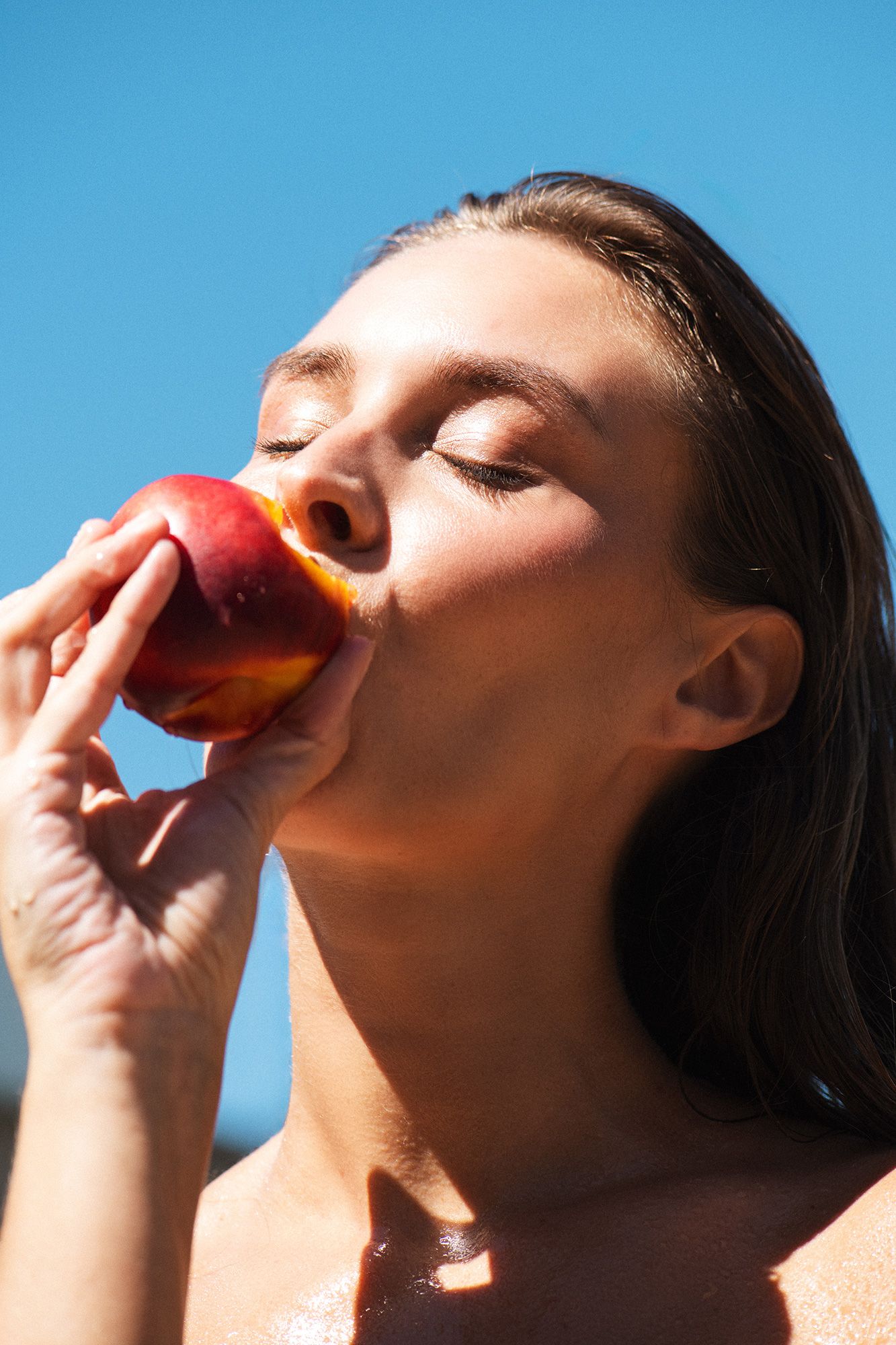 Young woman with closed eyes biting into a fresh ripe peach against a clear blue sky background.
