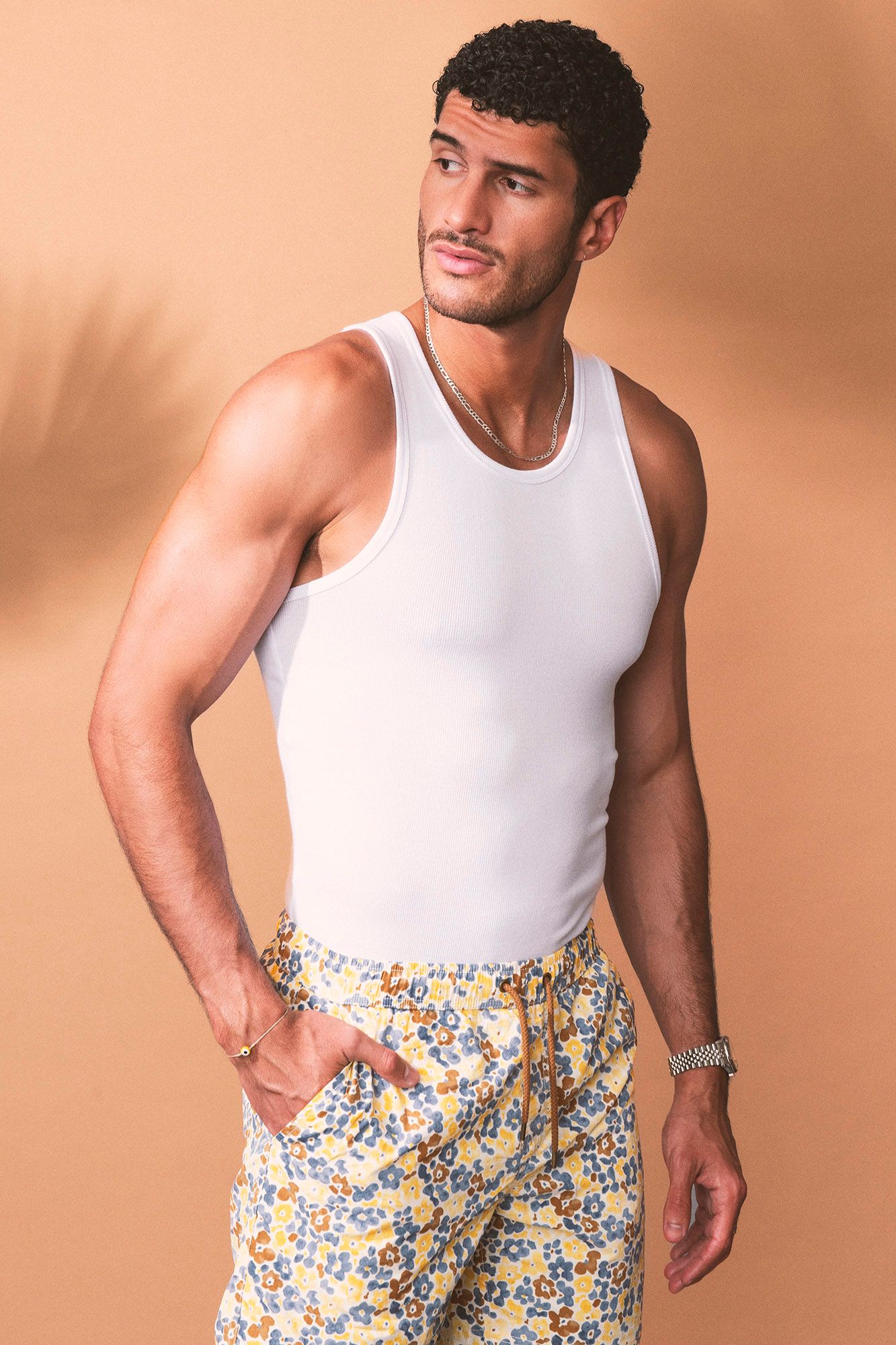 Man in white tank top and floral shorts posing against beige background.