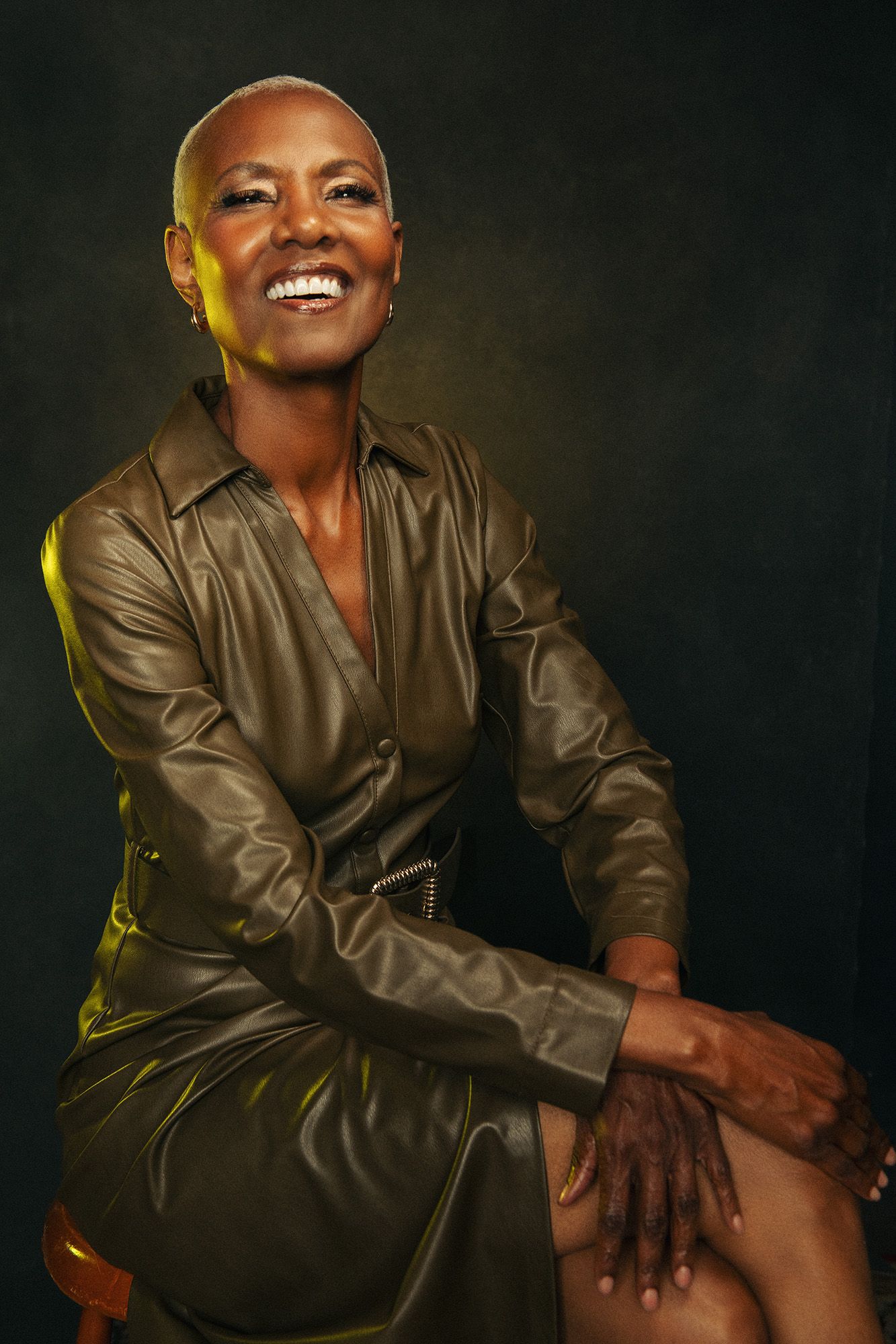 Smiling bald Black woman in olive leather dress seated confidently against dark studio background.