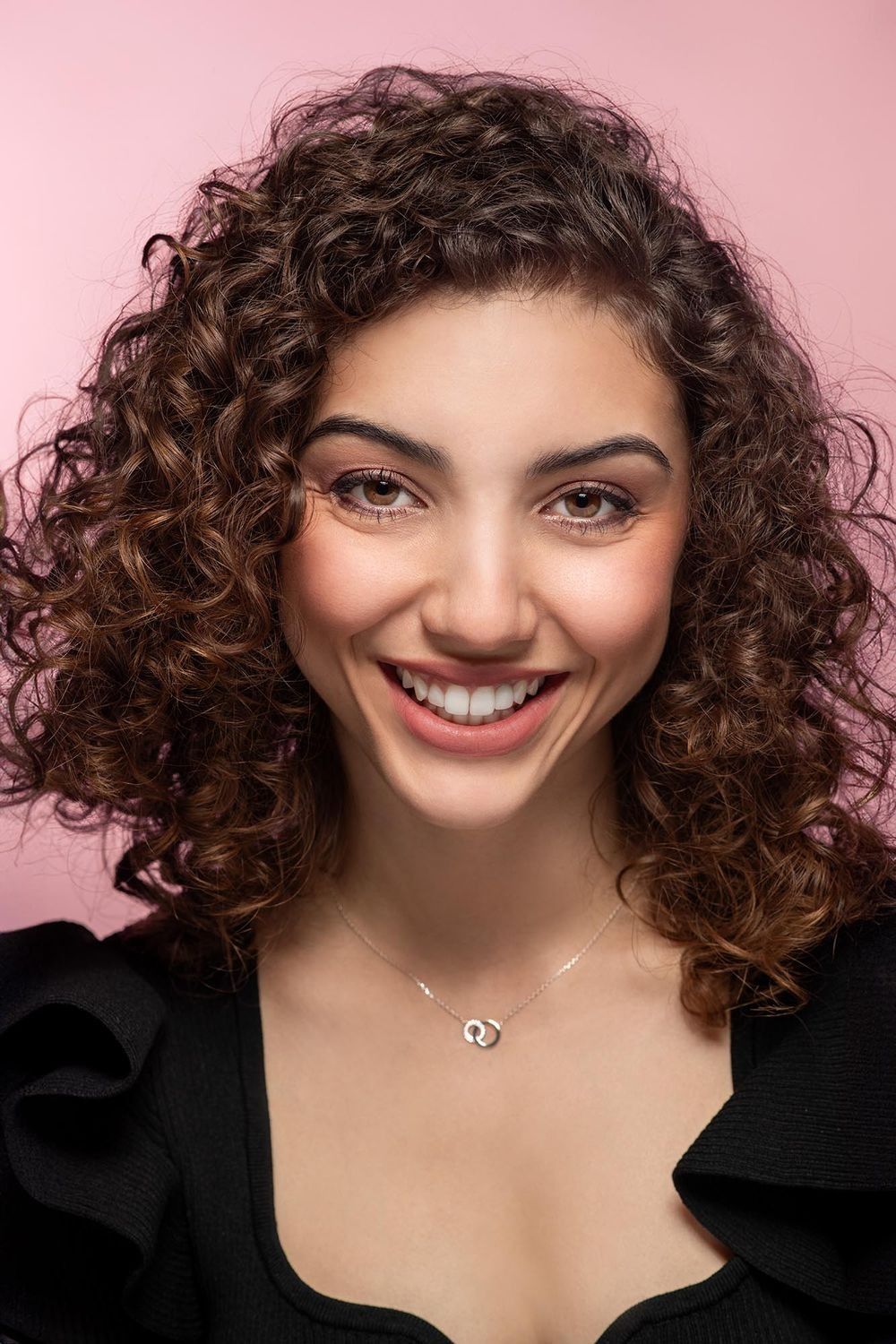 Smiling young woman with curly brown hair wearing a necklace against a pink background.