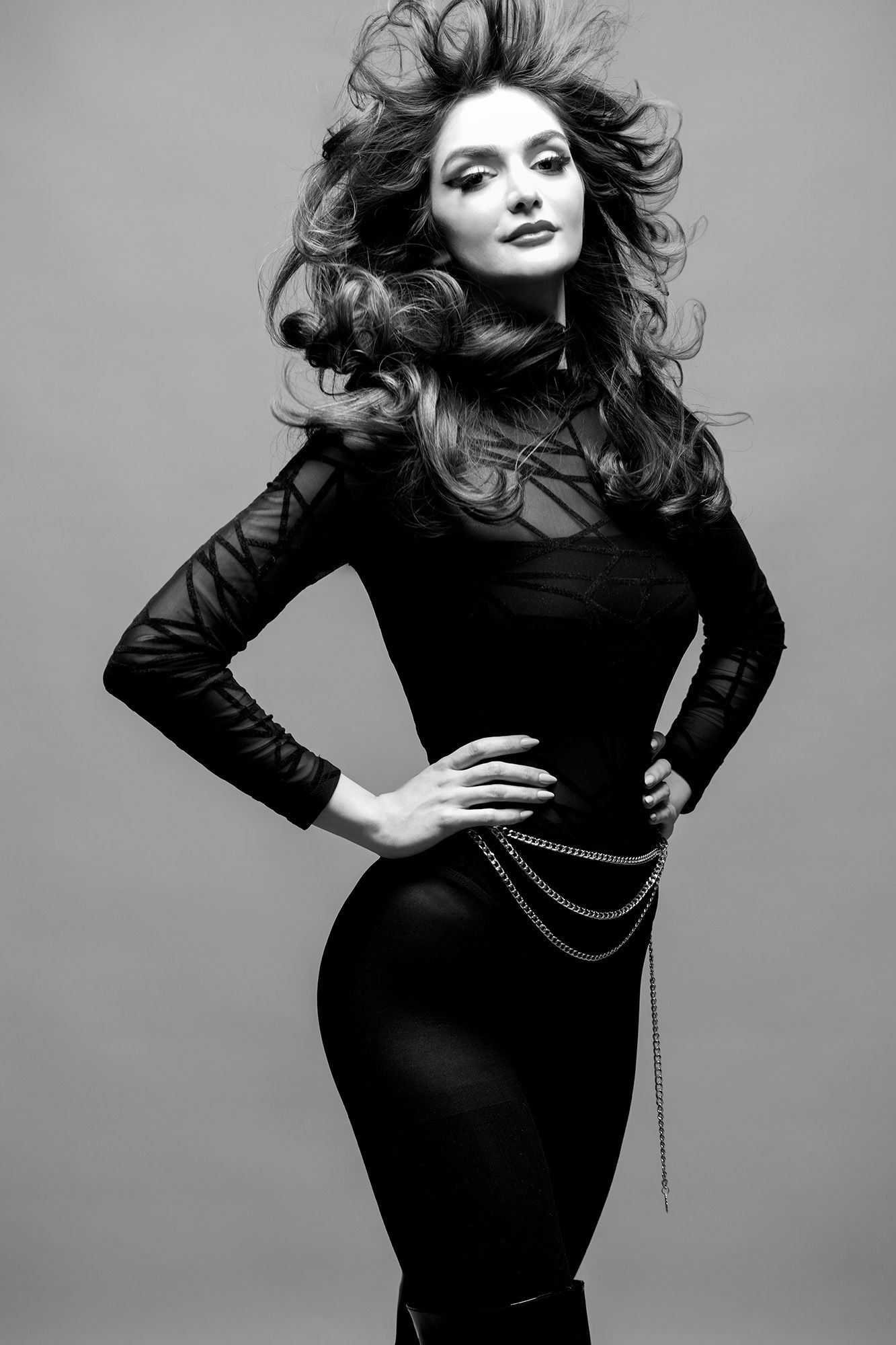 Elegant woman in black bodycon dress with chain belt and voluminous windswept hair in black and white photo.
