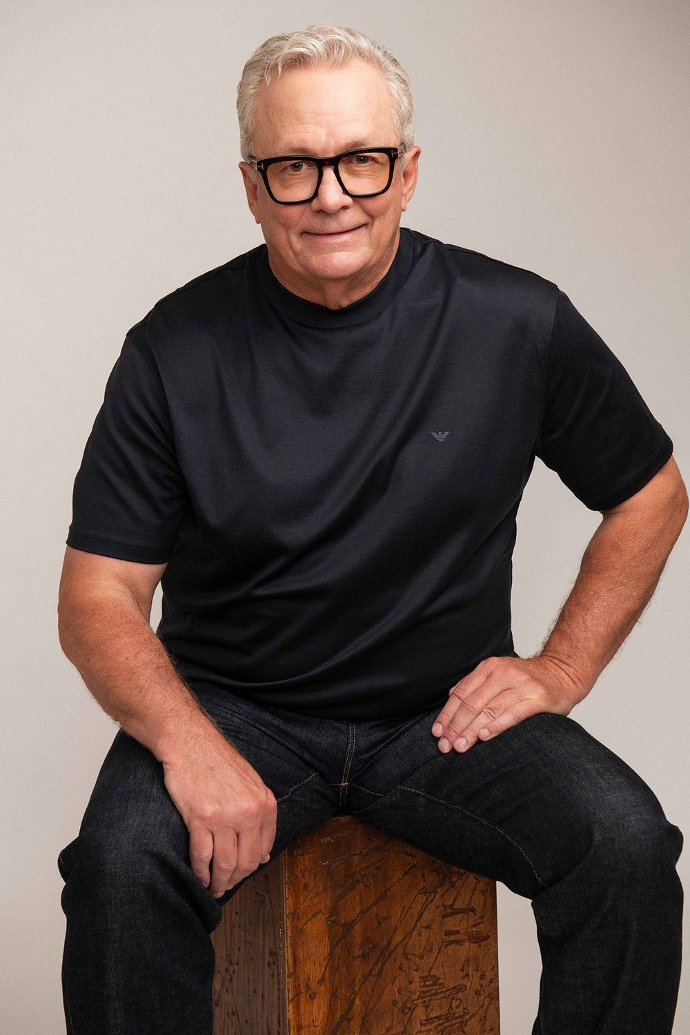 Mature man with gray hair and glasses sits casually in black Armani t-shirt and jeans.