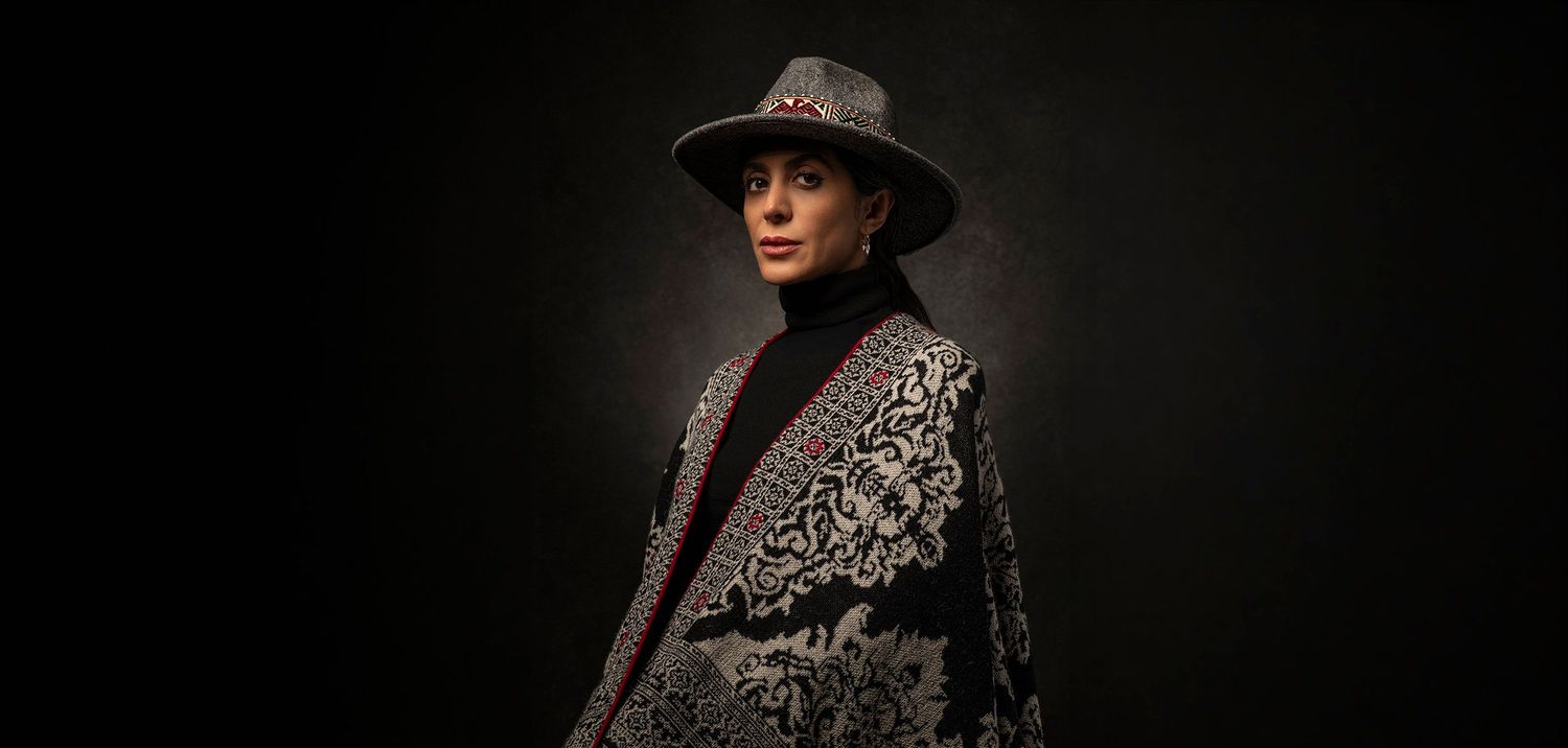 Stylish person in ornate black and white embroidered cape and wide-brim hat posing against dark background.