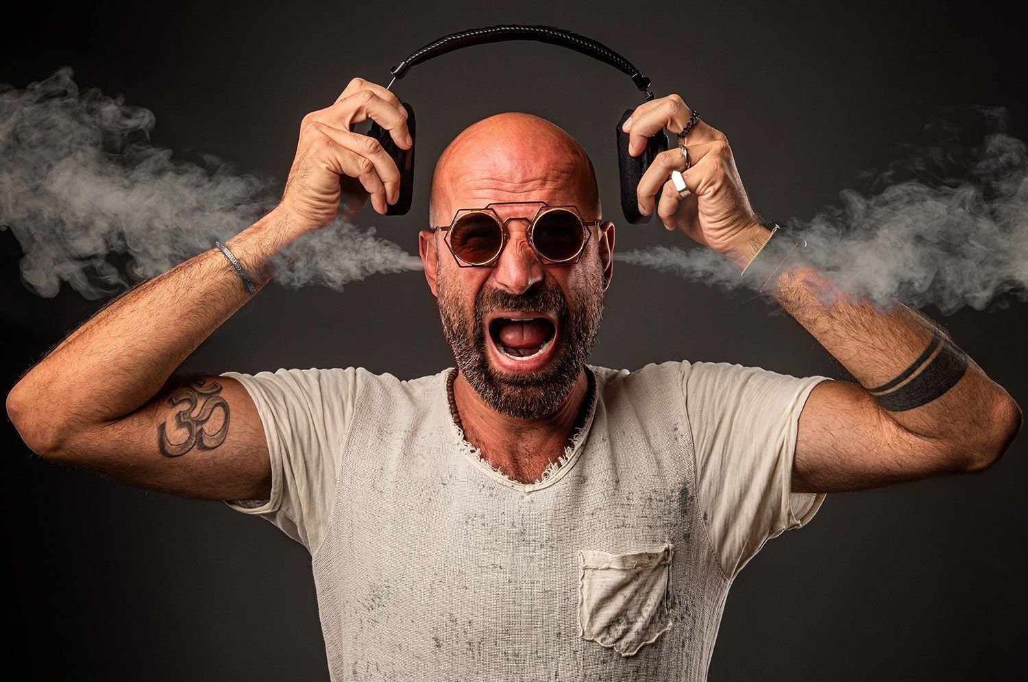 Bald man with tattoos screams while holding headphones with smoke effect on dark background.