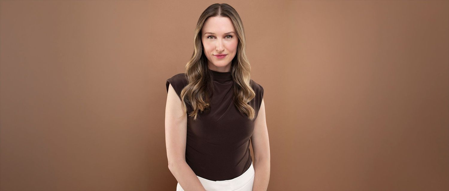 Woman in brown turtleneck top and white pants posing against warm brown background.