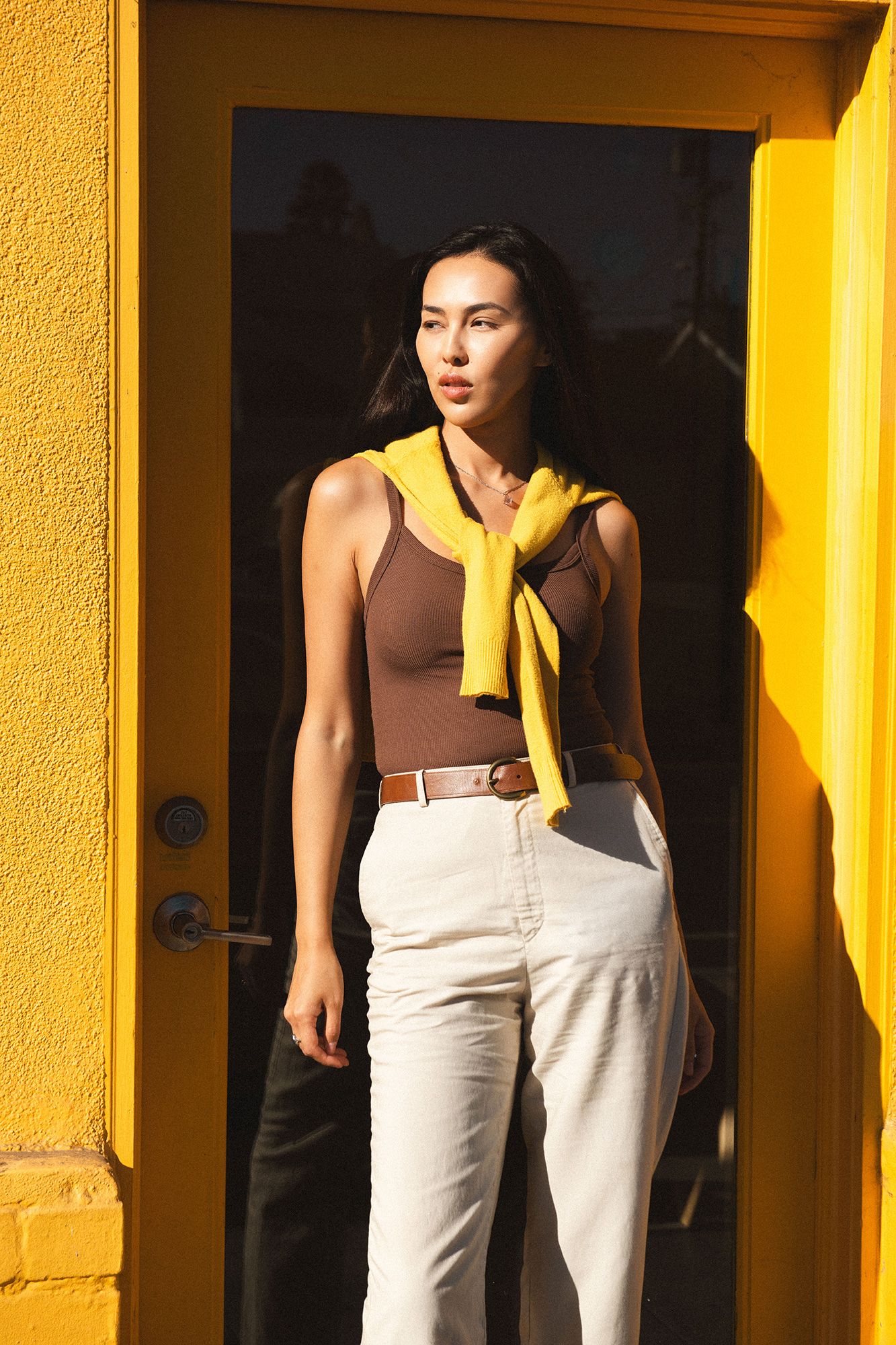 Woman in brown tank top, yellow scarf, and white pants standing by a yellow door in sunlight.