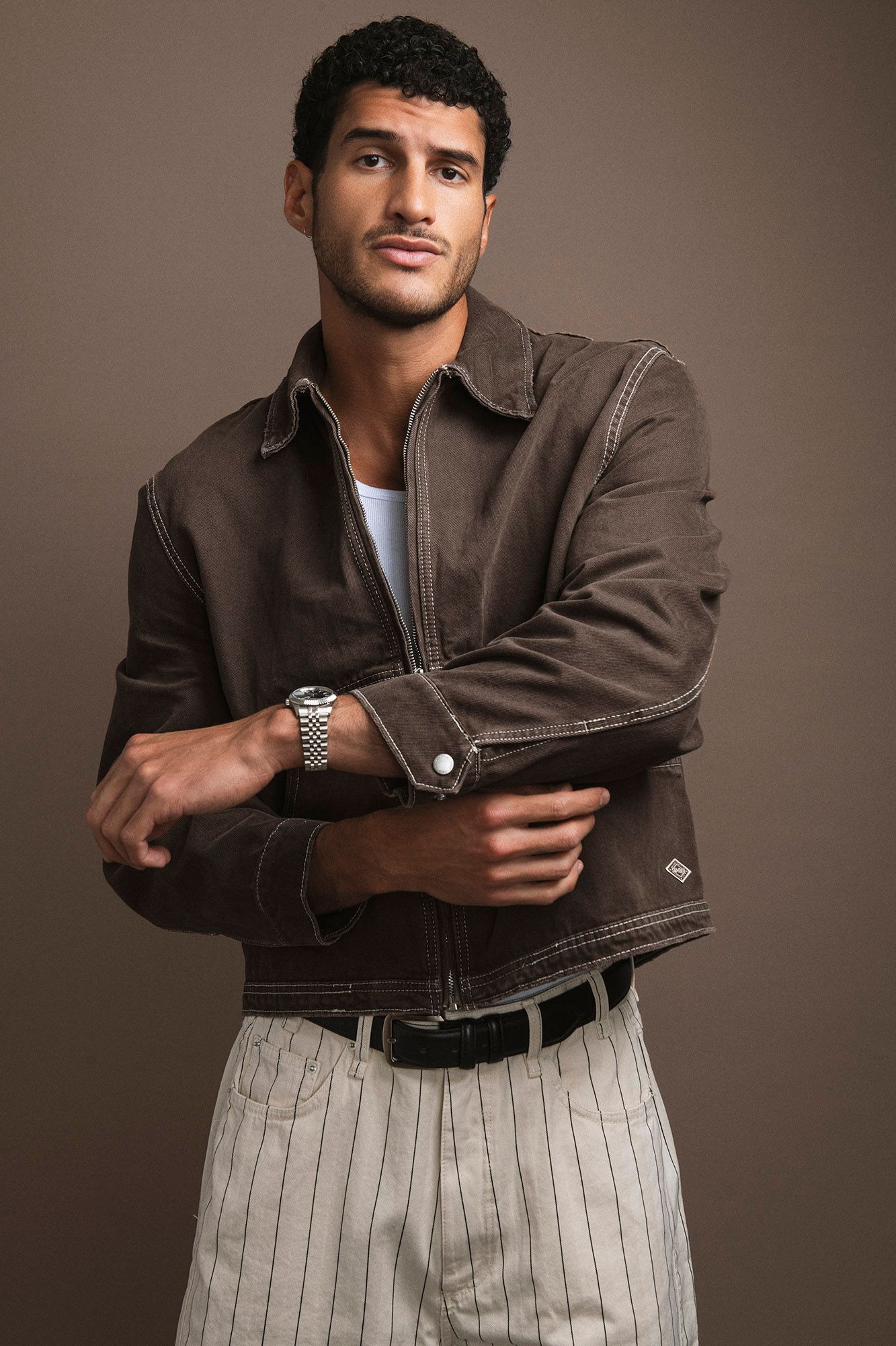 Man in brown jacket and striped trousers posing with arms crossed, wearing a silver watch.