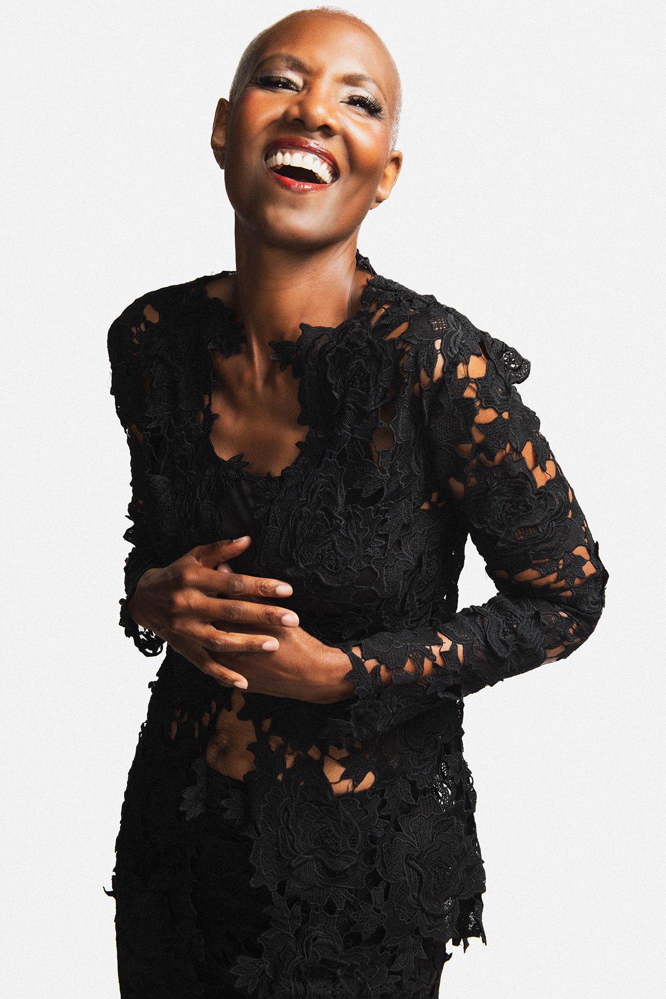 Laughing bald woman in elegant black lace dress posing confidently against white background.
