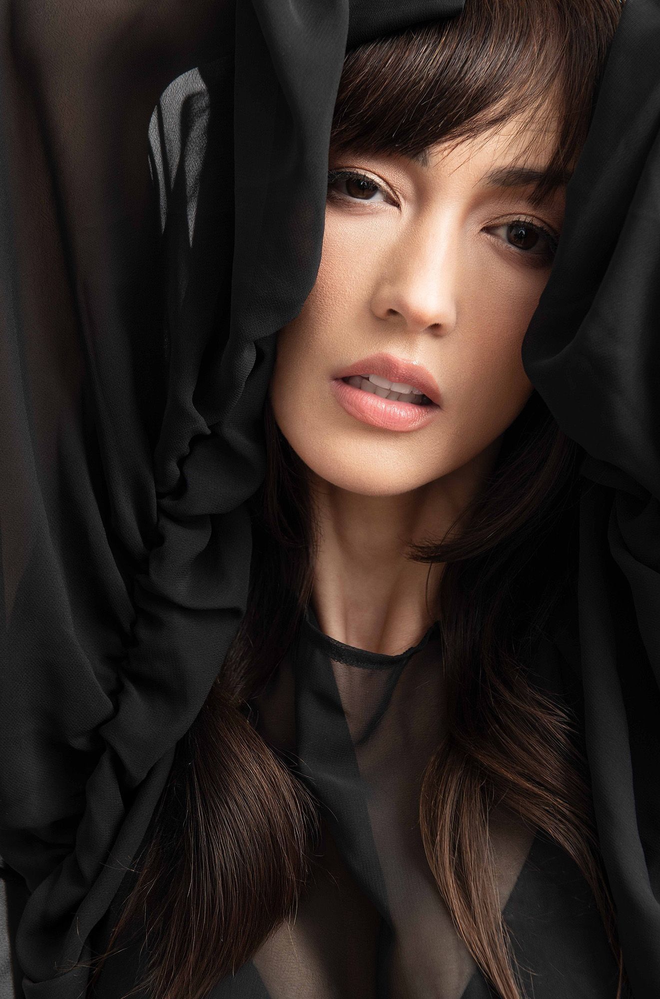 A woman with long brown hair peers through dramatic black fabric draping in a moody fashion portrait.