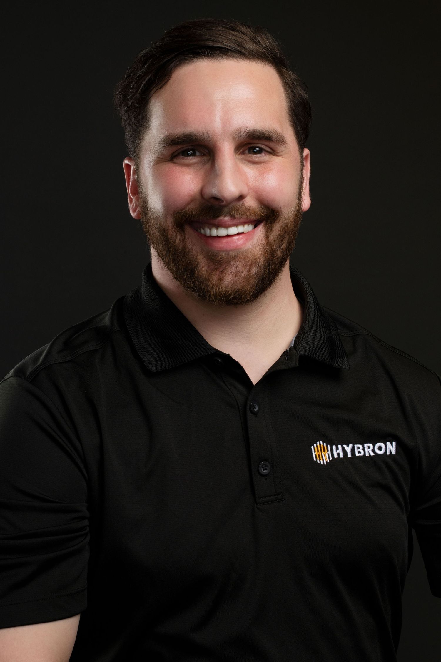 Smiling bearded man in black Hybron polo shirt against dark background.