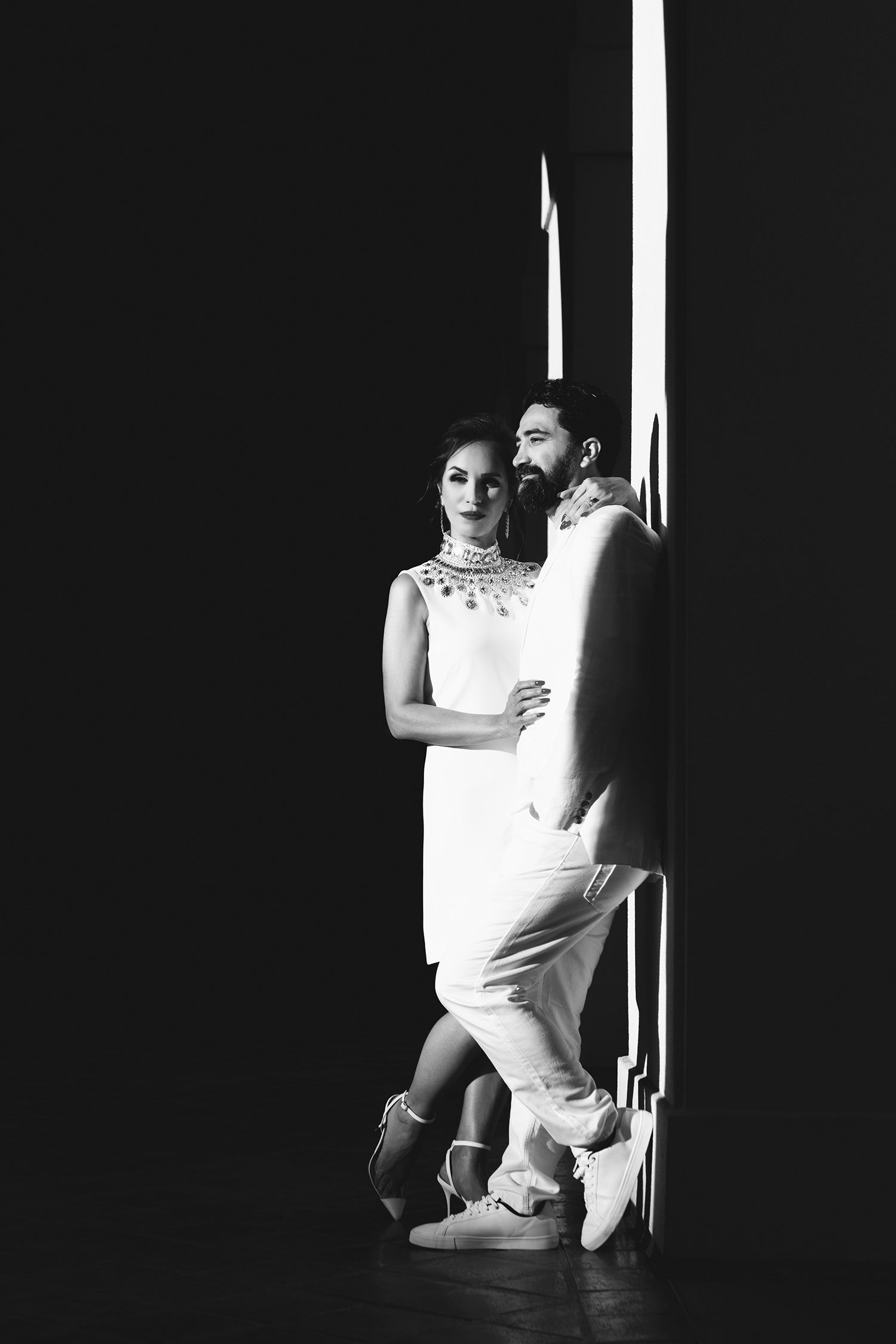 Elegant couple in white outfits pose in dramatic black and white portrait by a bright doorway.