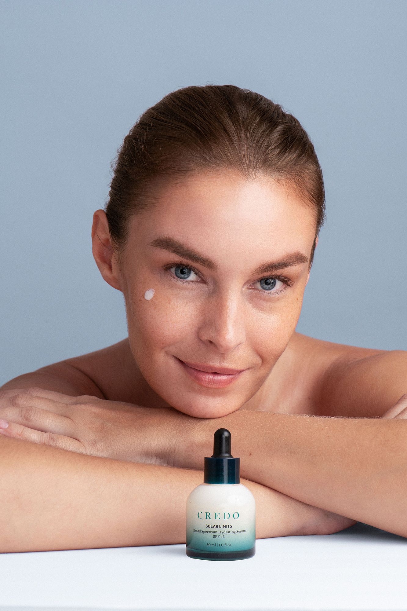 Woman posing with Credo beauty serum SPF dropper bottle, showcasing a dab of product on her cheek.
