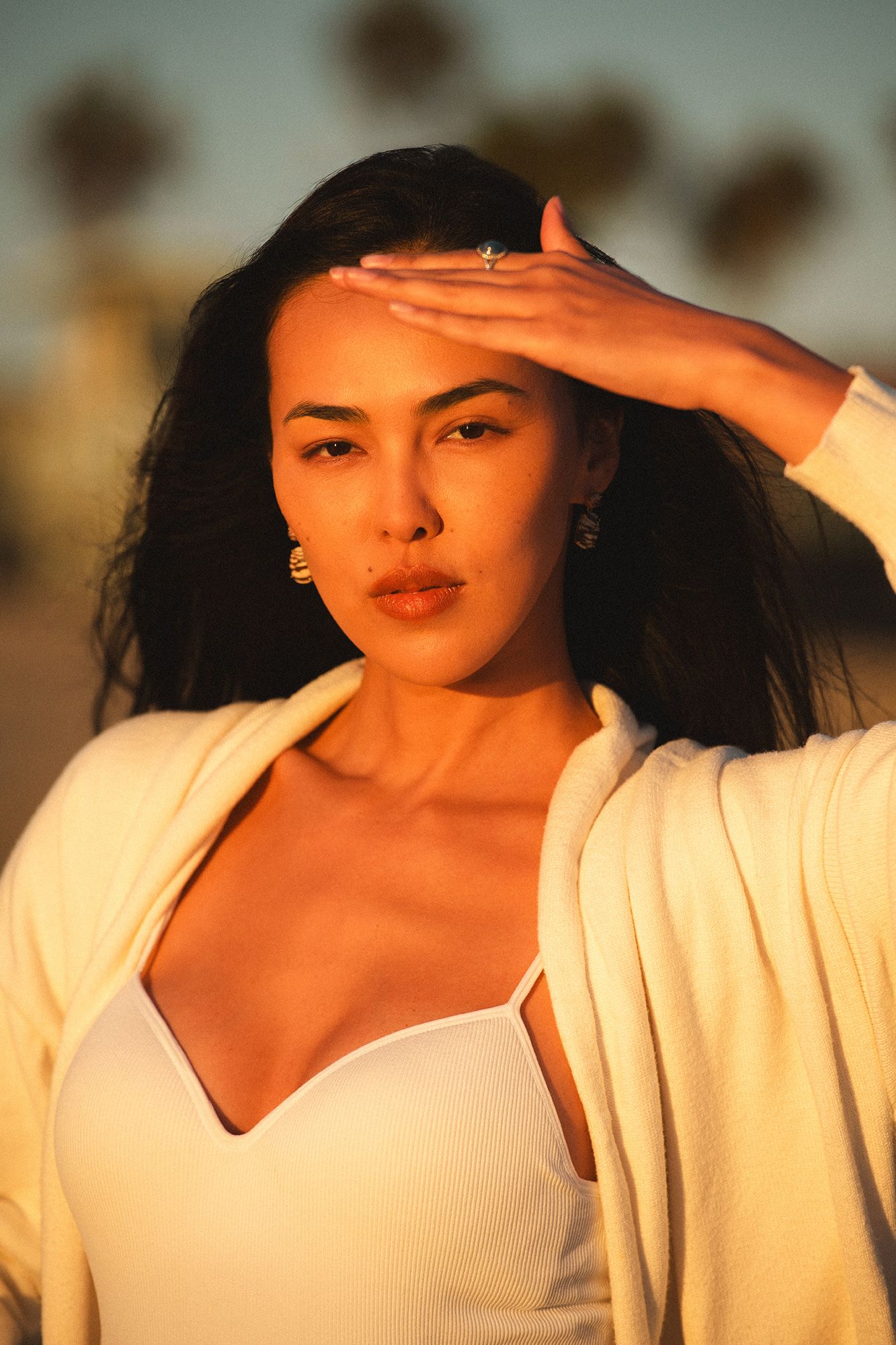 Woman shielding eyes from golden sunset light, wearing white outfit with palm trees in background.
