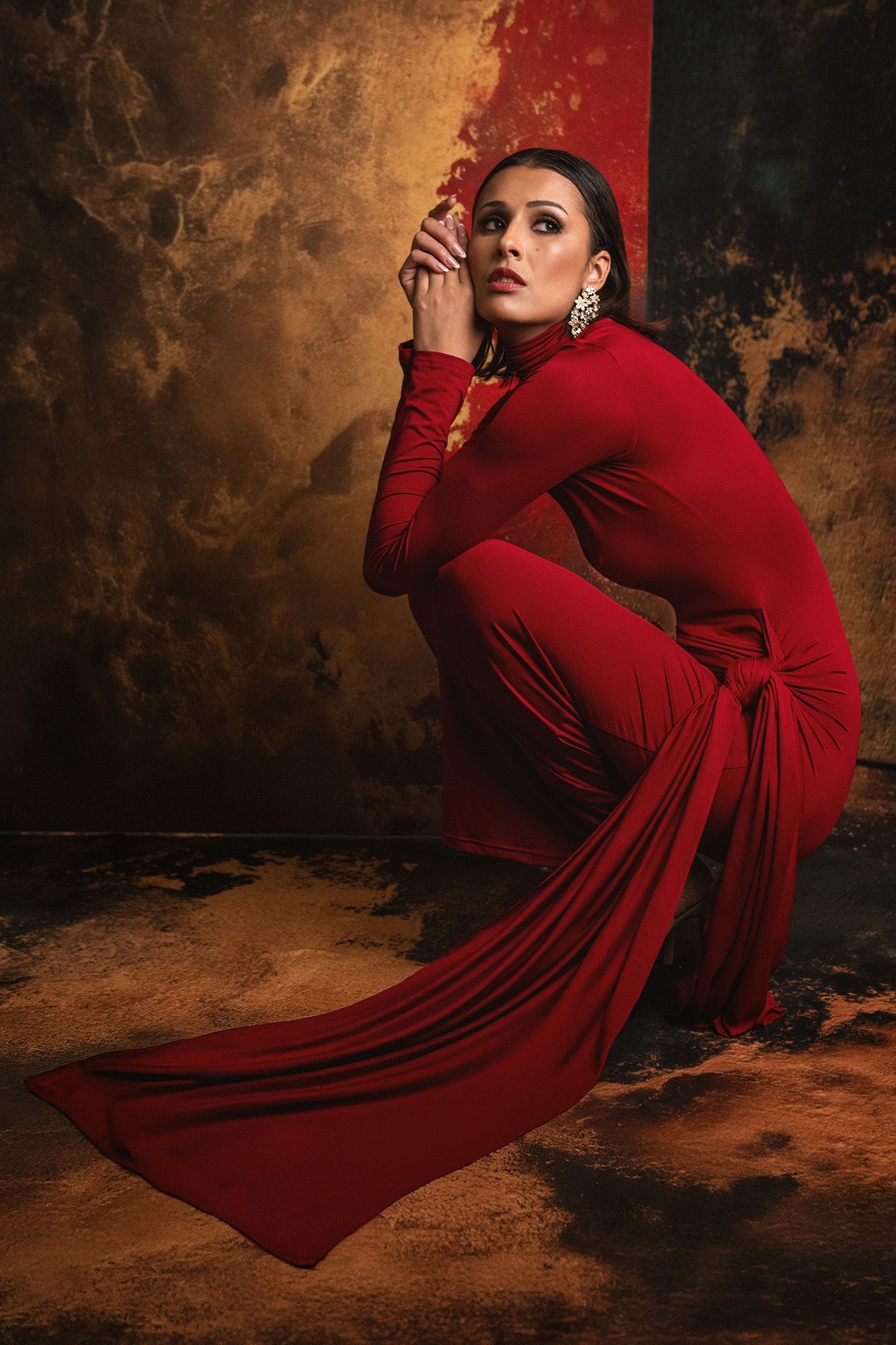 Woman in elegant red jumpsuit posing against warm textured backdrop.
