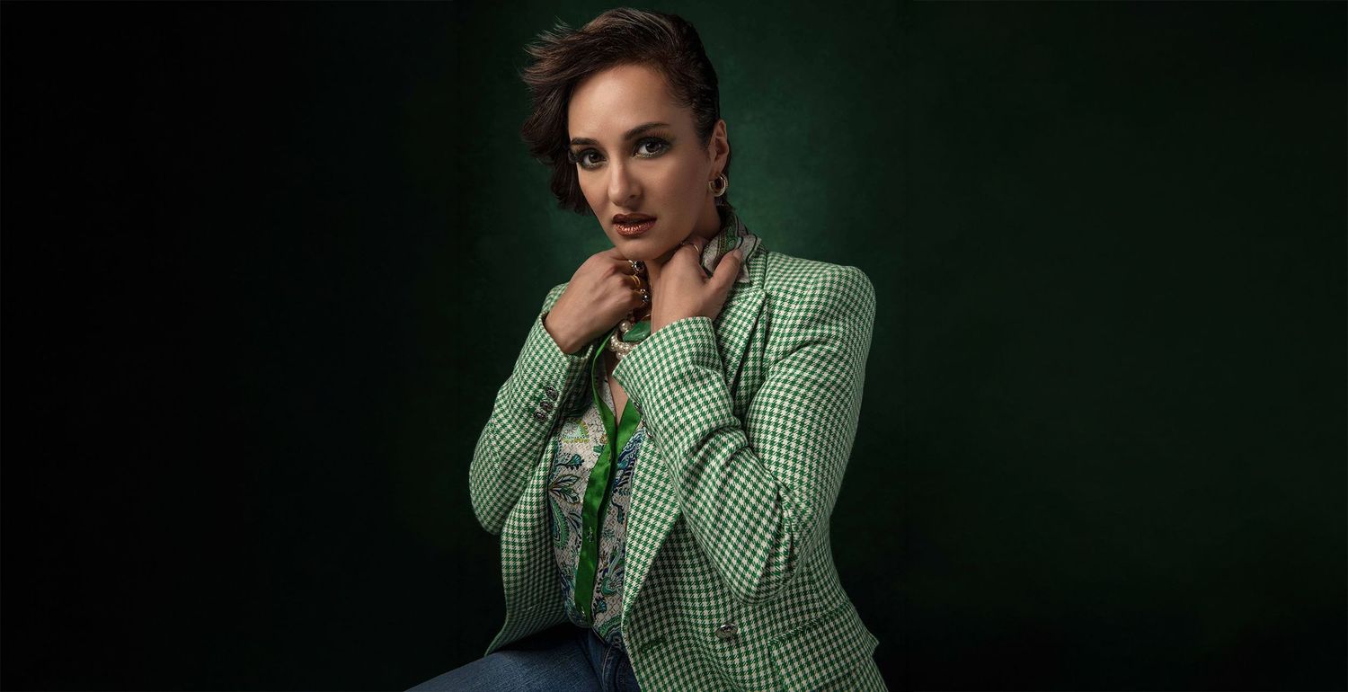 Woman in green houndstooth blazer posing confidently against dark green background in studio portrait.