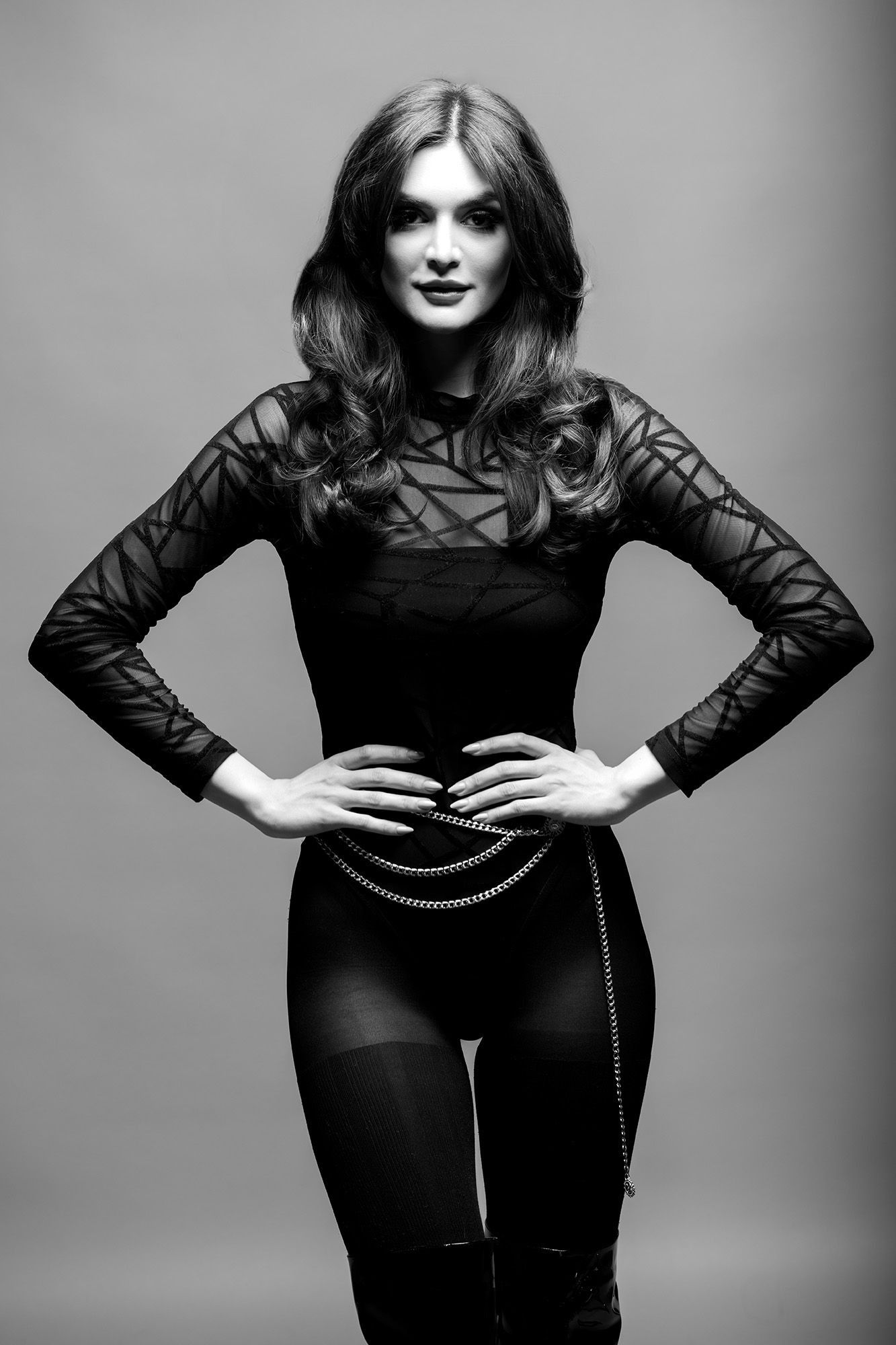 Stylish woman in black mesh bodysuit with chain belt posing with hands on hips in black and white photo.